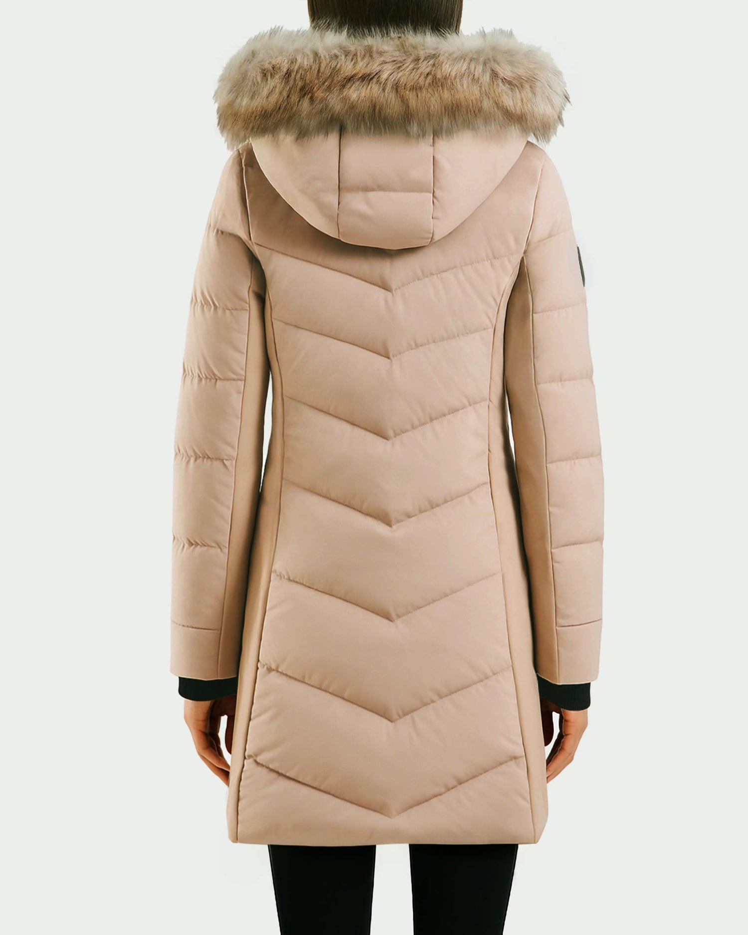 A person is seen from the back wearing the Rudsak LAUREN - 4124520 Taupe, a light beige knee-length quilted down coat with a water-repellent finish and faux fur hood, set against a plain background.