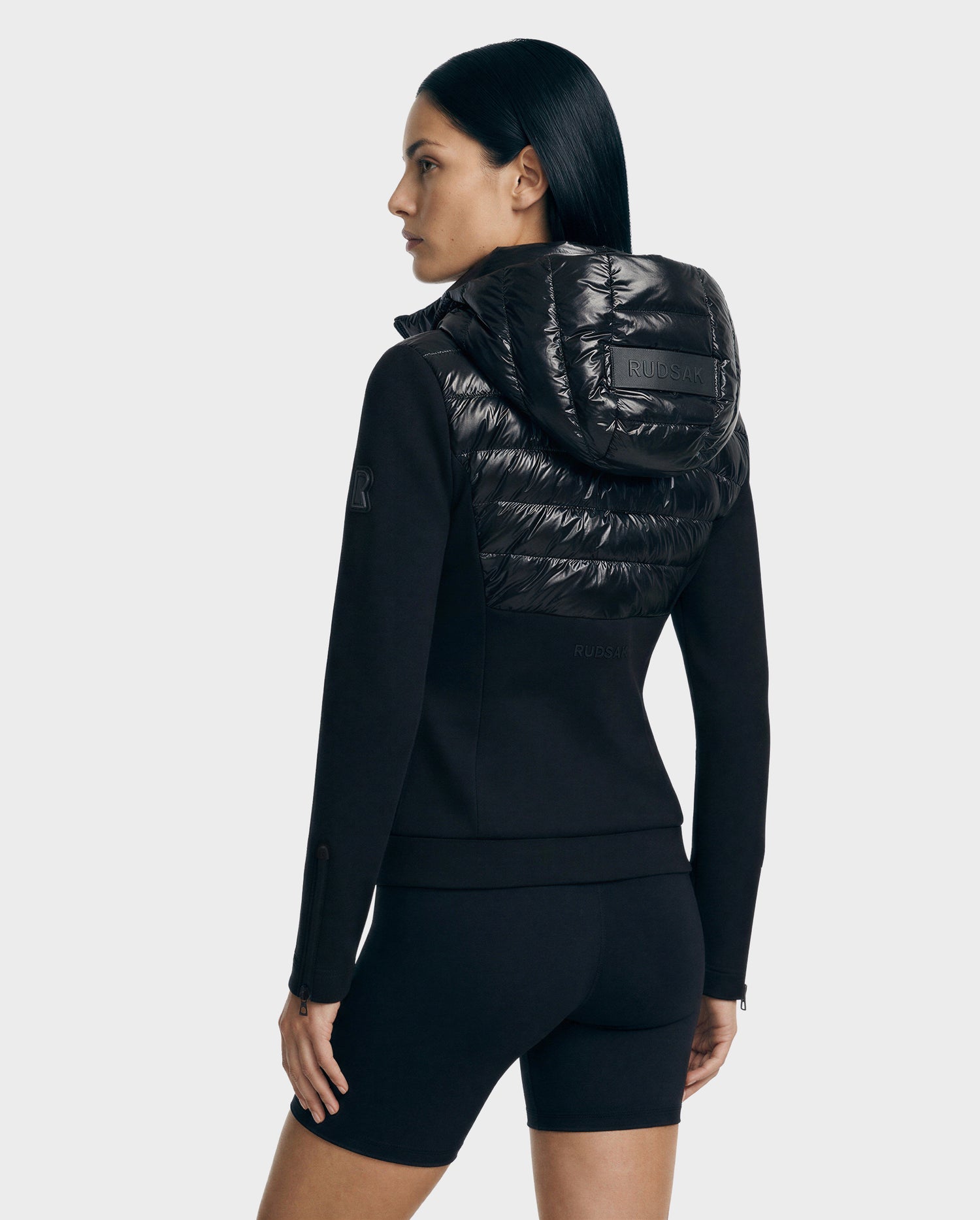 A woman with straight dark hair models the Rudsak MABS C - 8125701 Black fitted jacket featuring a shiny, quilted hood and matching black shorts, standing slightly turned against a plain light background.