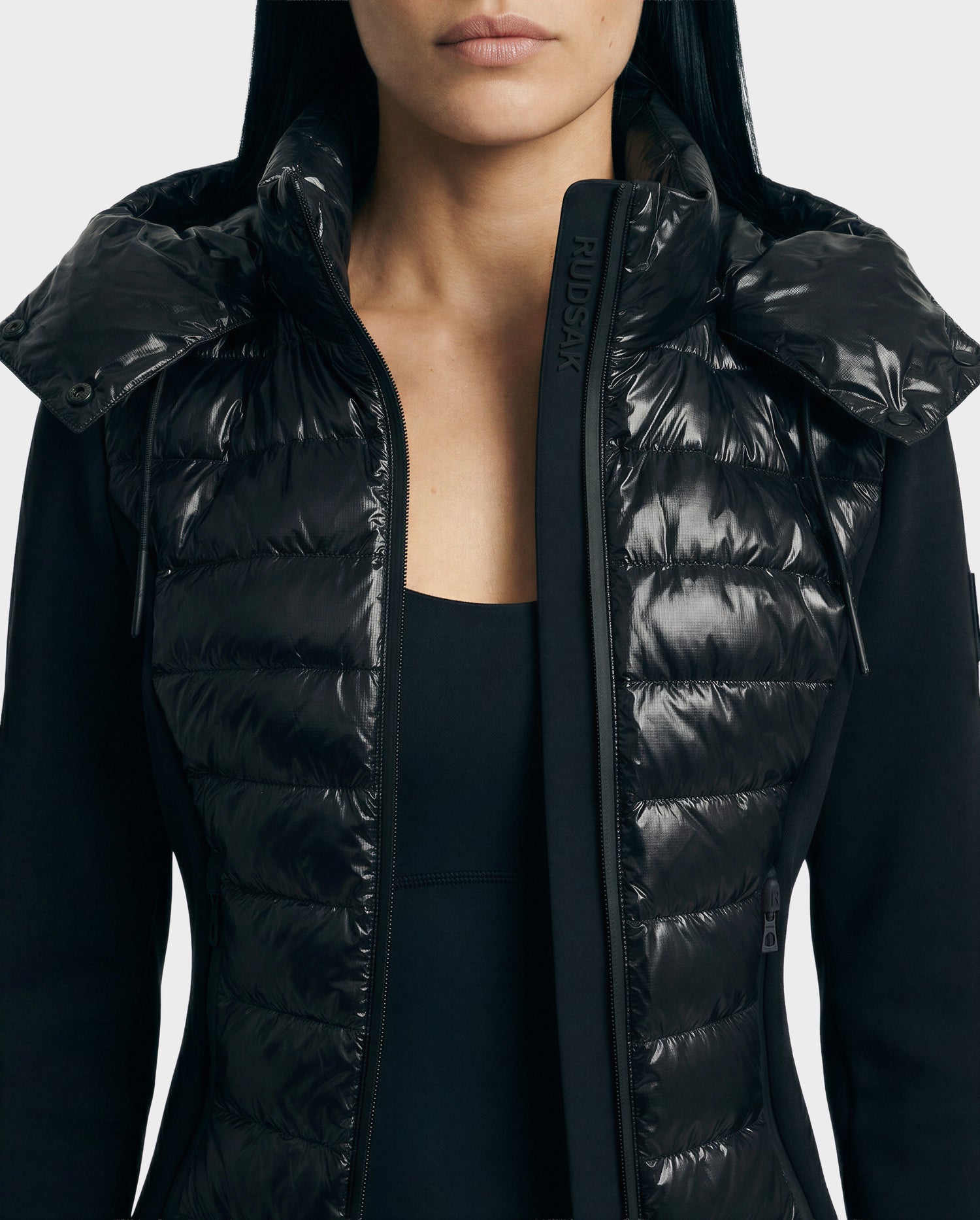 A woman wears the Rudsak MABS C - 8125701 Black puffer jacket with the collar open, revealing a black top. The image is cropped below her nose.