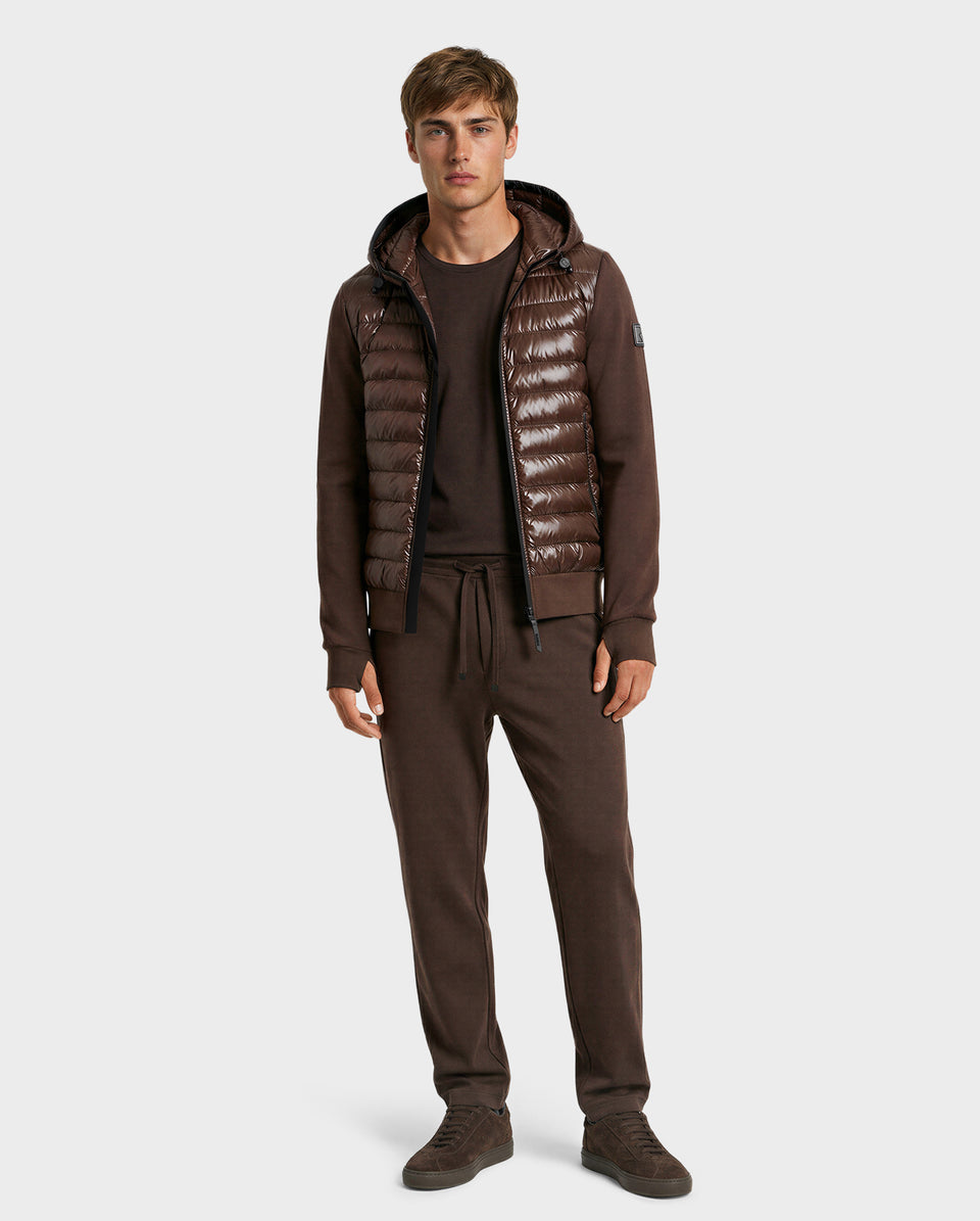 A man faces forward in the Rudsak DEE S - 6125684 Espresso men’s lightweight down hooded jacket over a brown sweater and joggers, paired with brown sneakers—perfect for urban style—against a simple, light backdrop.