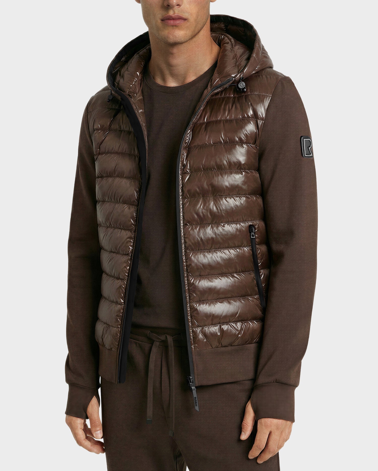 A man wears the Rudsak DEE S - 6125684 Espresso, a shiny brown men’s lightweight down jacket with a hood, over matching brown long-sleeve shirt and pants, standing against a plain light background.