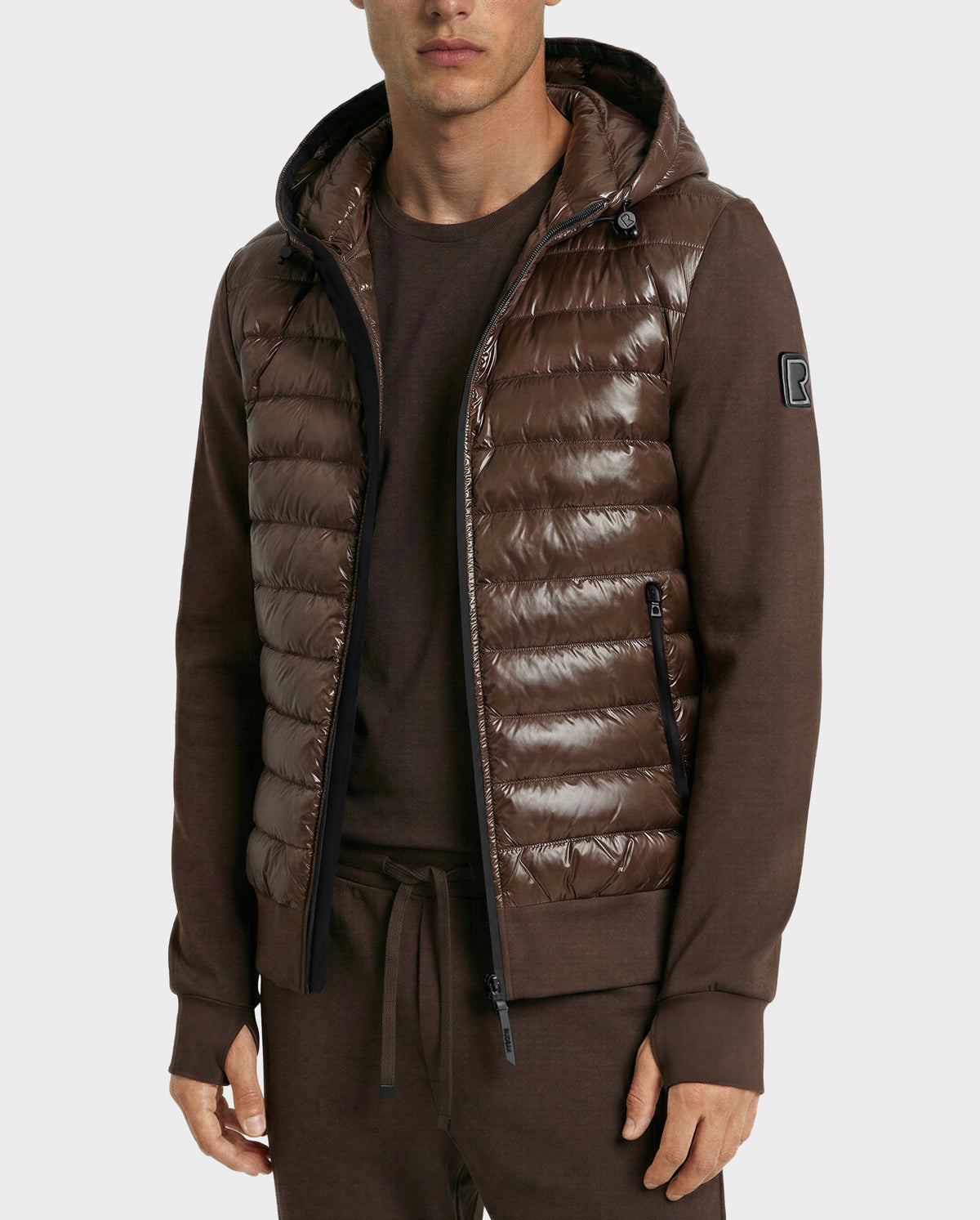 A man wears the Rudsak DEE S - 6125684 Espresso, a shiny brown men’s lightweight down jacket with a hood, over matching brown long-sleeve shirt and pants, standing against a plain light background.