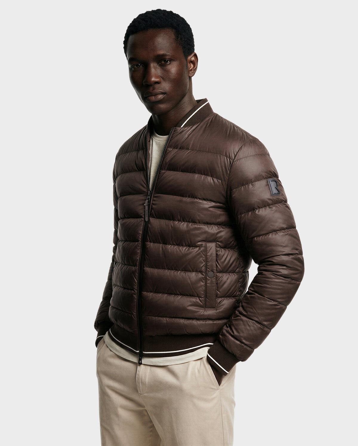 A man wears the Rudsak JEREMY - 6125025 Espresso men’s down bomber jacket over a light sweater and beige pants, standing with hands in pockets against a plain light background.