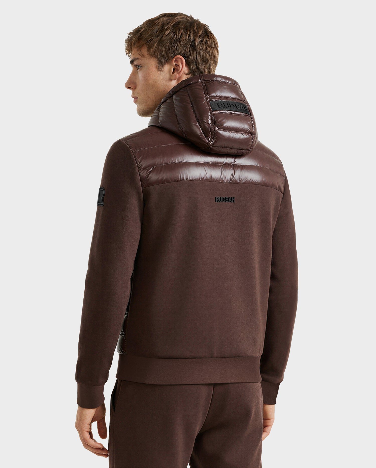 A person is shown from the back wearing the Rudsak DEE S - 6125684 Espresso hoodie with a shiny, padded hood and shoulders, filled with RDS-certified duck down, paired with matching brown pants and logos on the hood and upper back.