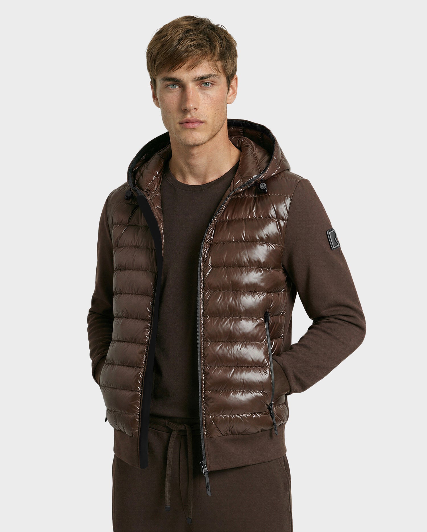 A young man with light brown hair models the Rudsak DEE S - 6125684 Espresso men’s lightweight down jacket, made from RDS-certified duck down, layered over a matching brown outfit, standing hands-in-pockets against a plain light background.