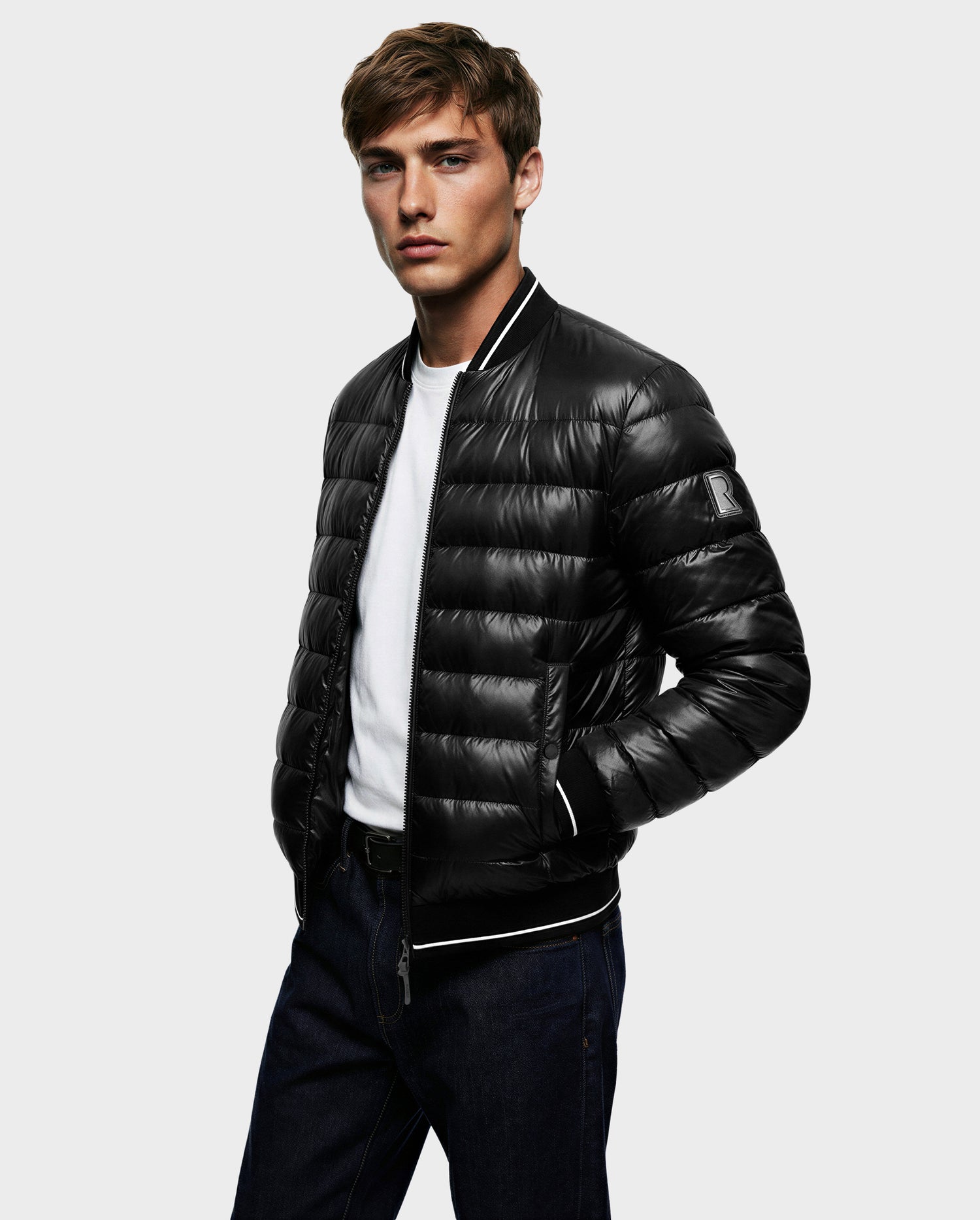A young man wears the Rudsak JEREMY - 6125025 Black men’s down bomber jacket over a white T-shirt and dark jeans, standing against a plain light gray background with his hands in his jacket pockets, looking at the camera.