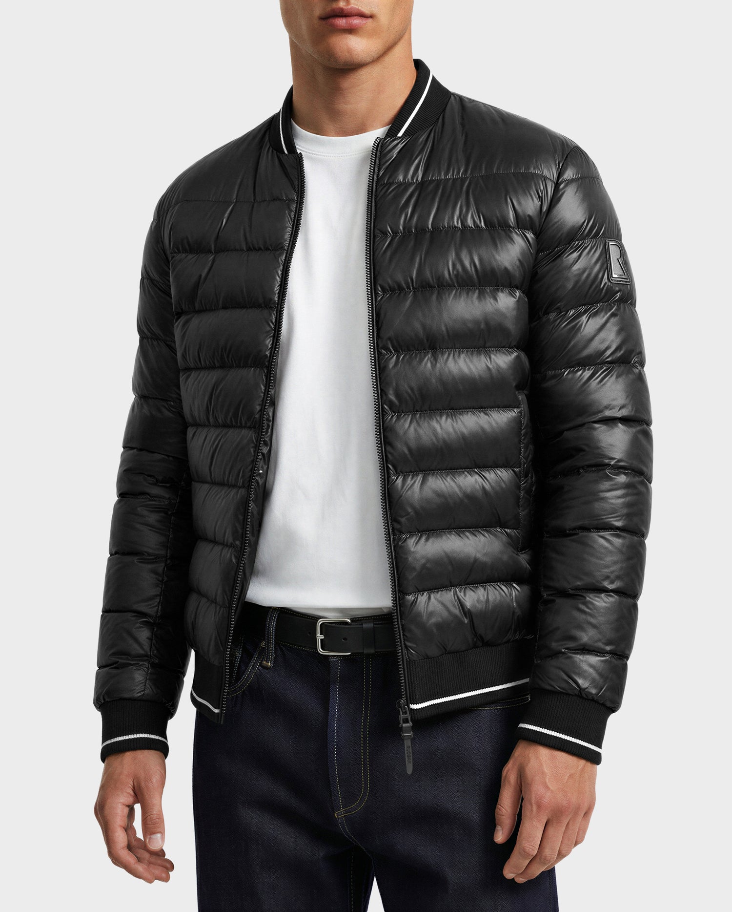 A man wearing the Rudsak JEREMY - 6125025 Black down bomber jacket, a white t-shirt, dark jeans, and a black belt stands against a plain light background.