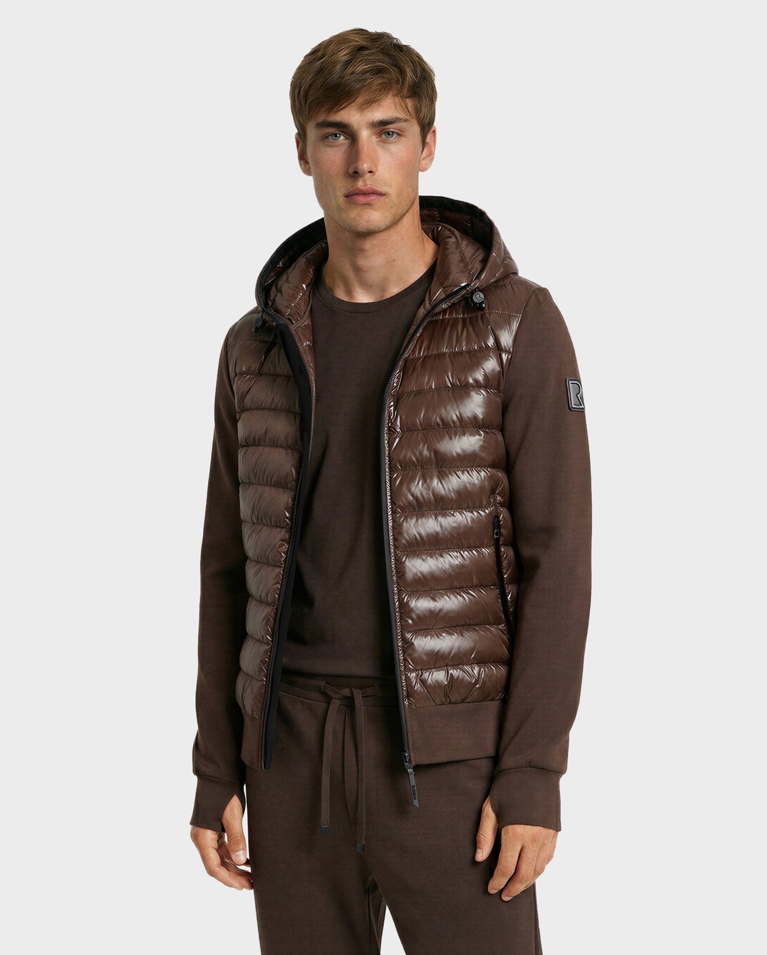 A young man stands facing the camera against a plain light background, wearing the Rudsak DEE S - 6125684 Espresso, a shiny brown men’s lightweight down jacket with knit sleeves, paired with matching brown pants and shirt.
