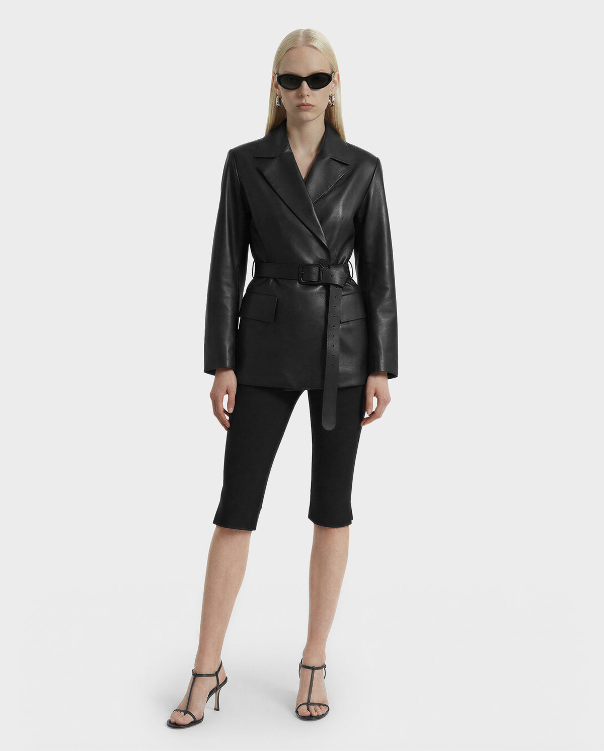 A woman wearing the Rudsak KENDALL - 8125617 Black tailored blazer, styled as a leather jacket, with black knee-length shorts, strappy black heels, sunglasses, and drop earrings stands facing forward against a plain light background.