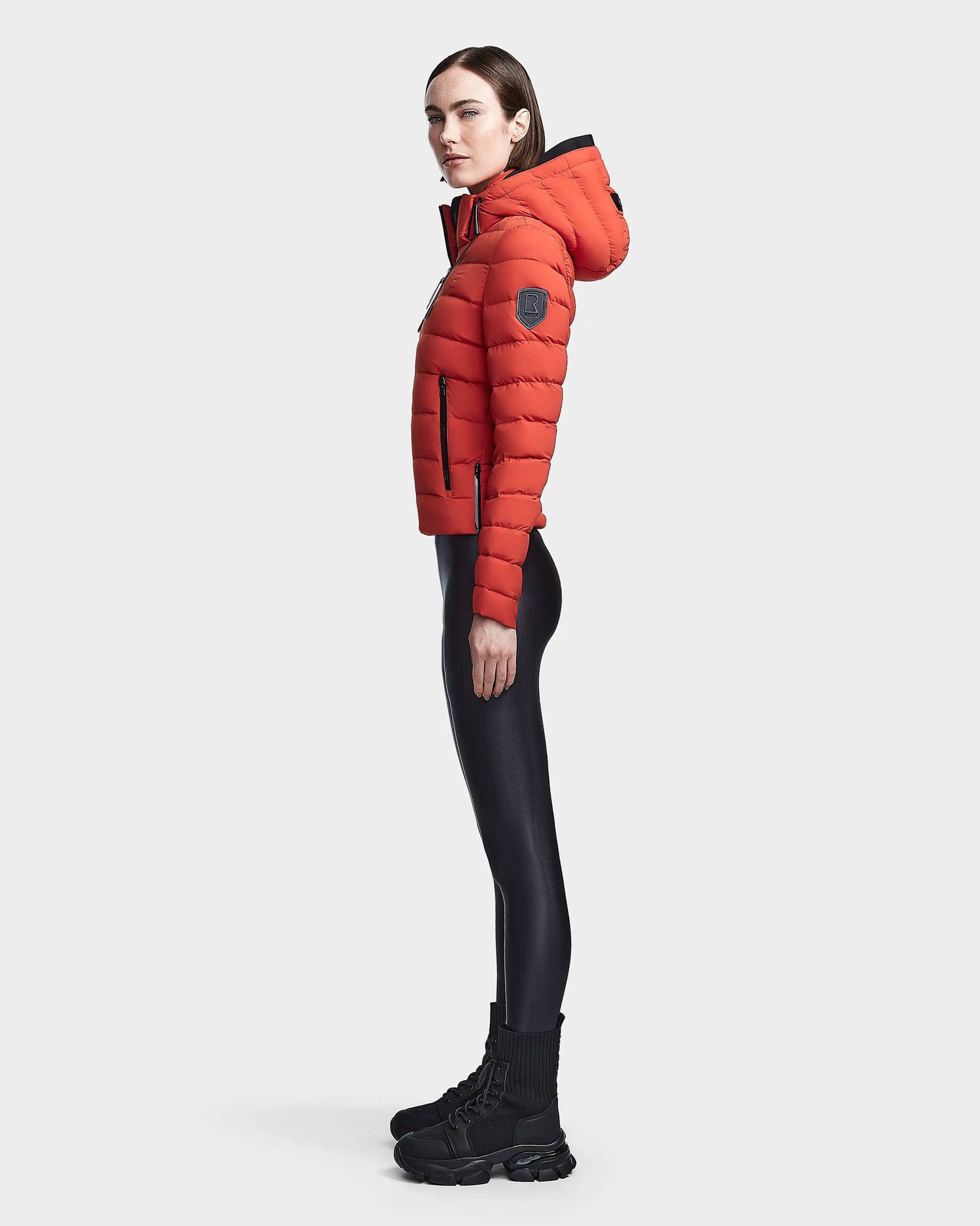 A woman stands in profile wearing the Rudsak MELISSA - 8122551 Orange puffer jacket with a hood, paired with black shiny leggings and chunky black boots against a plain white background.