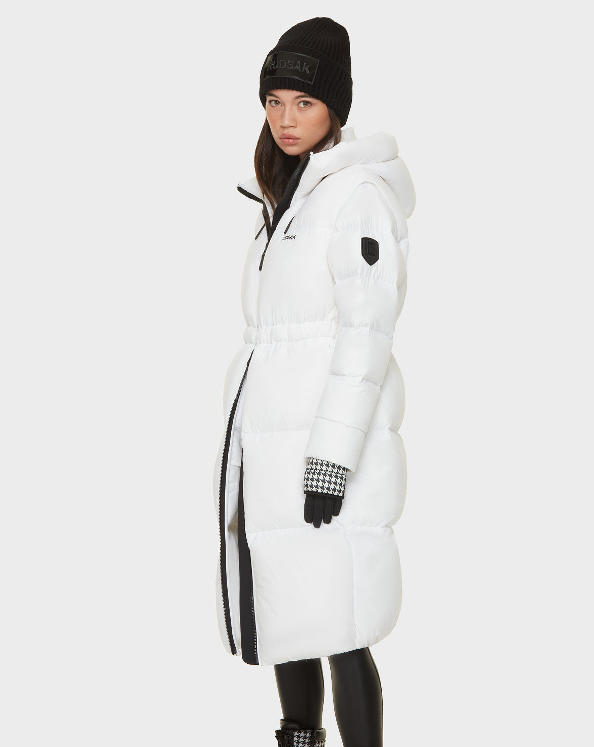 A woman in the Rudsak MATILA - 8121537 OPTIC WHITE long down puffer coat with hood, black knit beanie, gloves, and leggings stands turned slightly to the side against a plain white background.