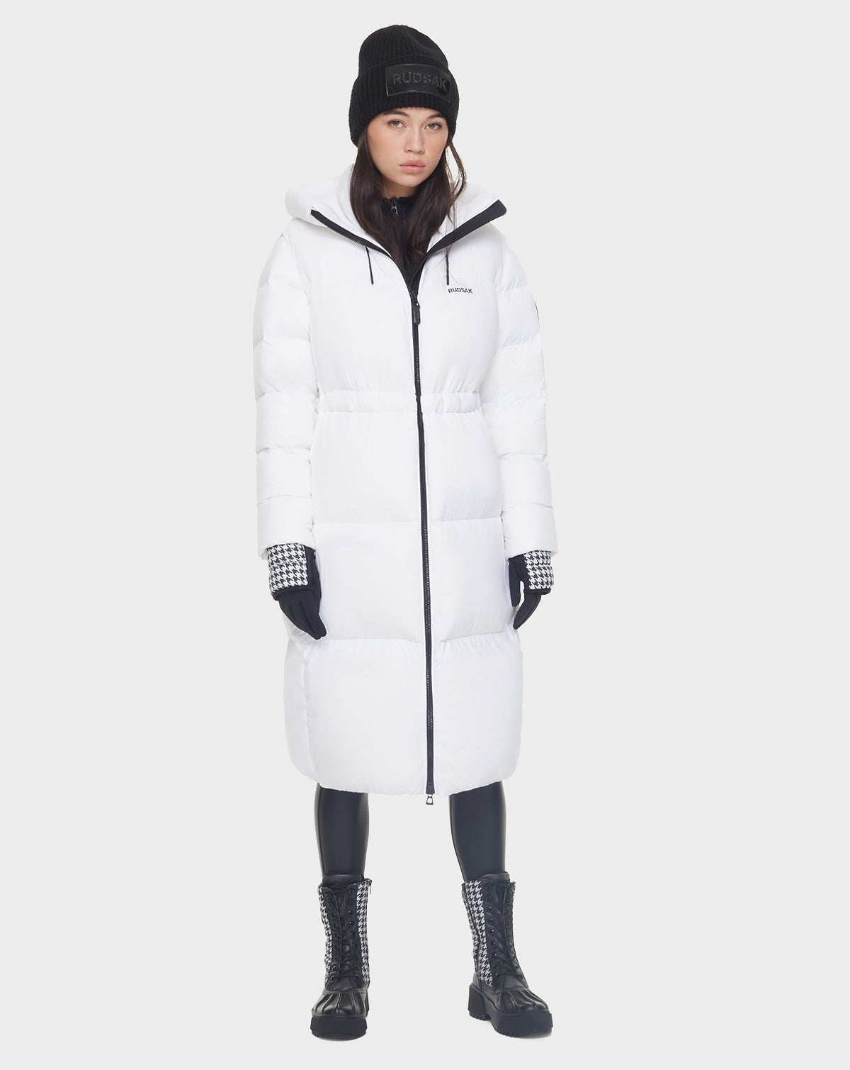 A woman in the Rudsak MATILA - 8121537 OPTIC WHITE down puffer coat, black beanie, houndstooth-cuffed black gloves, black leggings, and lace-up boots stands against a plain light background.