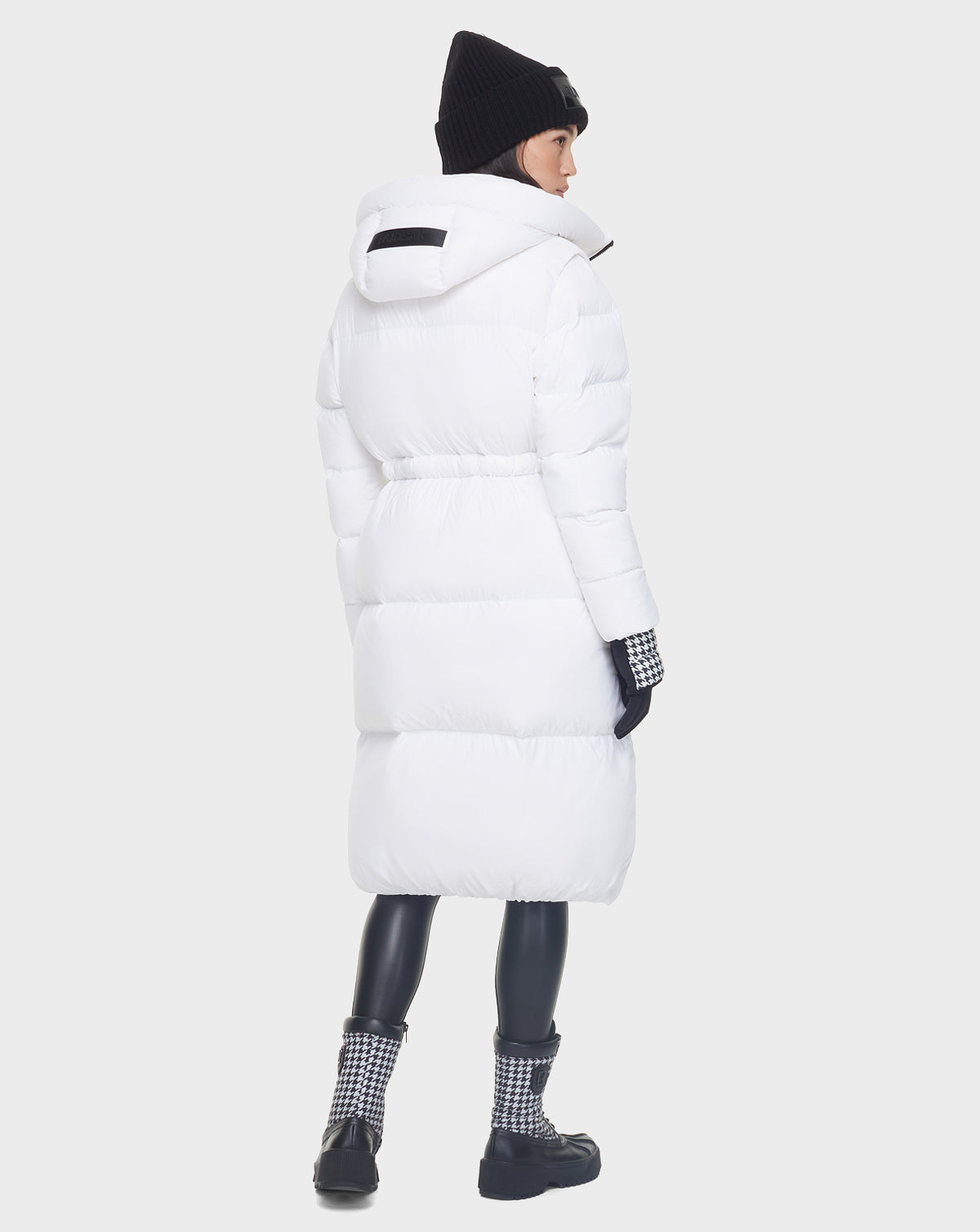 A person faces away, showcasing the back of the Rudsak MATILA - 8121537 OPTIC WHITE women’s down puffer coat with RDS-certified down, paired with a black beanie, gloves, leggings, and patterned boots against a plain light background.