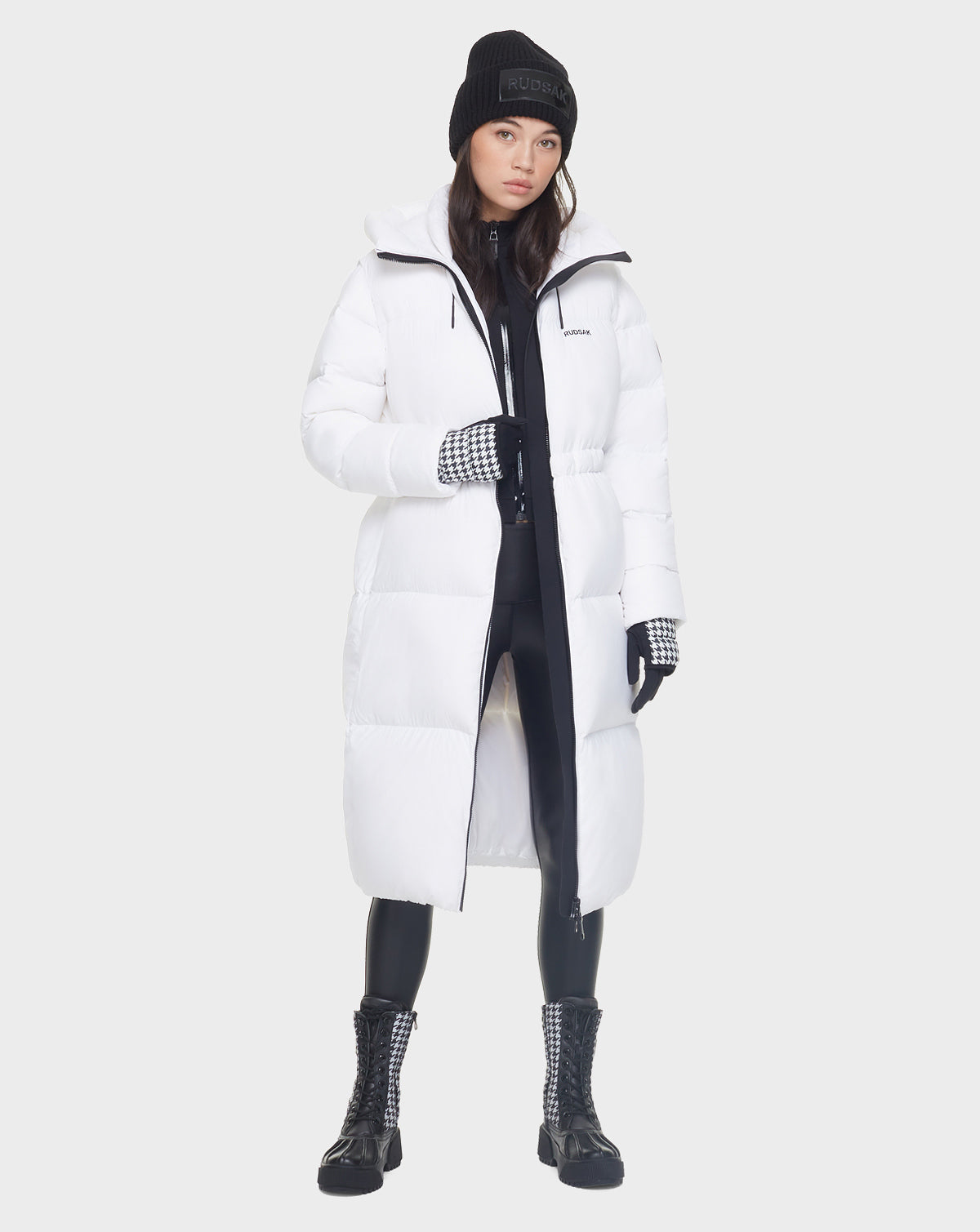 A woman in the Rudsak MATILA - 8121537 OPTIC WHITE long down puffer coat, black leggings, lace-up boots, gloves, and a beanie stands against a plain light background.
