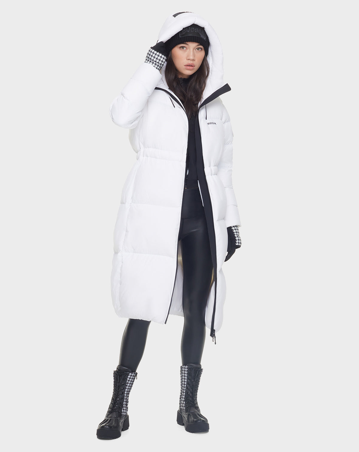 A woman in the Rudsak MATILA - 8121537 OPTIC WHITE long puffer coat stands against a plain light background, styled with black leggings, black combat boots, a black knit hat, and checkered gloves.