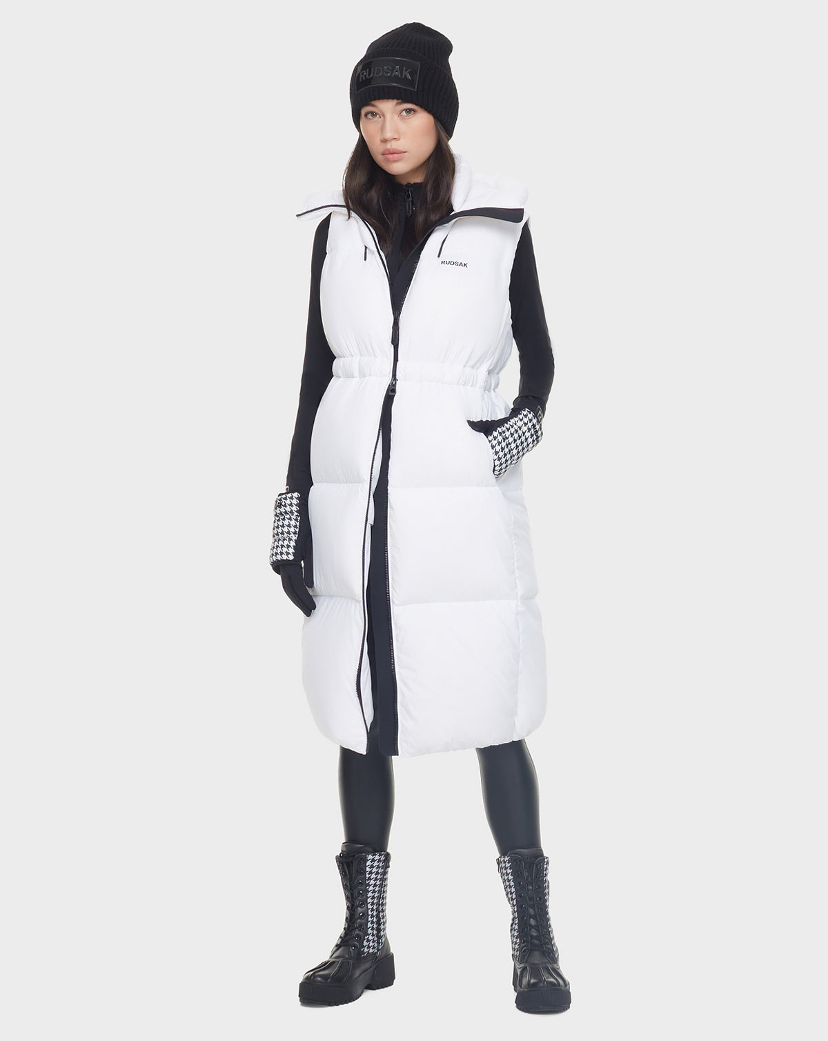 A woman wears the Rudsak MATILA - 8121537 OPTIC WHITE long down puffer vest over a black turtleneck, leggings, boots, checkered gloves, and a black beanie, standing against a plain light gray background.