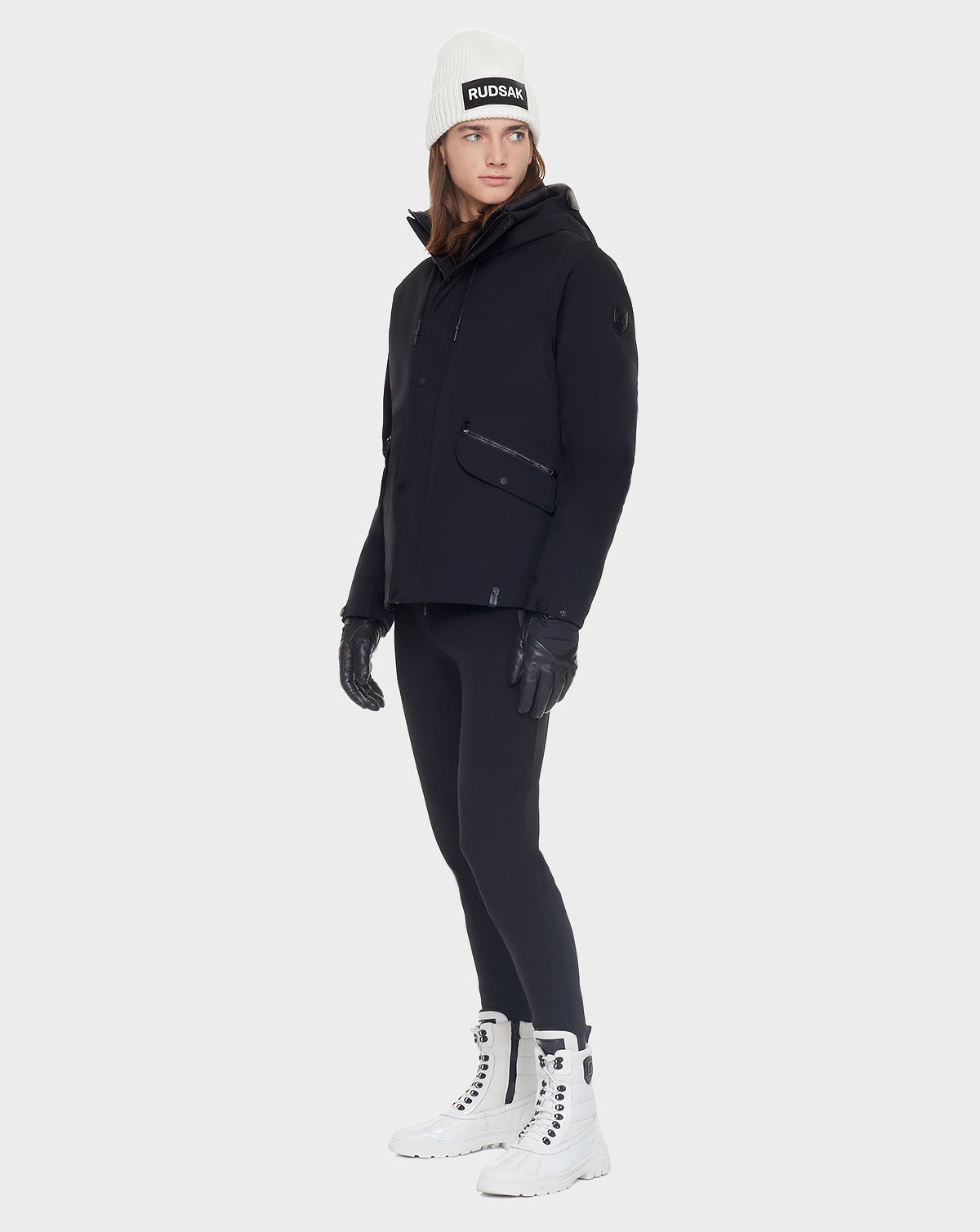 A person wearing the Rudsak DEST - CA EN 6119516 BLACK goose down hooded jacket, black leggings, gloves, a white knit beanie, and white lace-up boots stands against a plain light background.