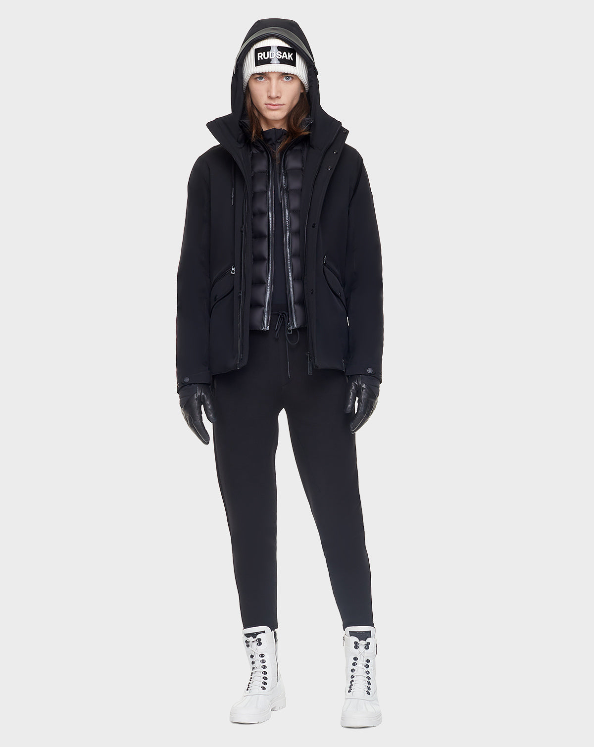 A person stands facing forward, wearing the Rudsak DEST - CA EN 6119516 BLACK hooded down jacket over a puffer vest, black gloves, black pants, white high-top sneakers, and a white RUBAIYAT beanie. The background is plain light gray.