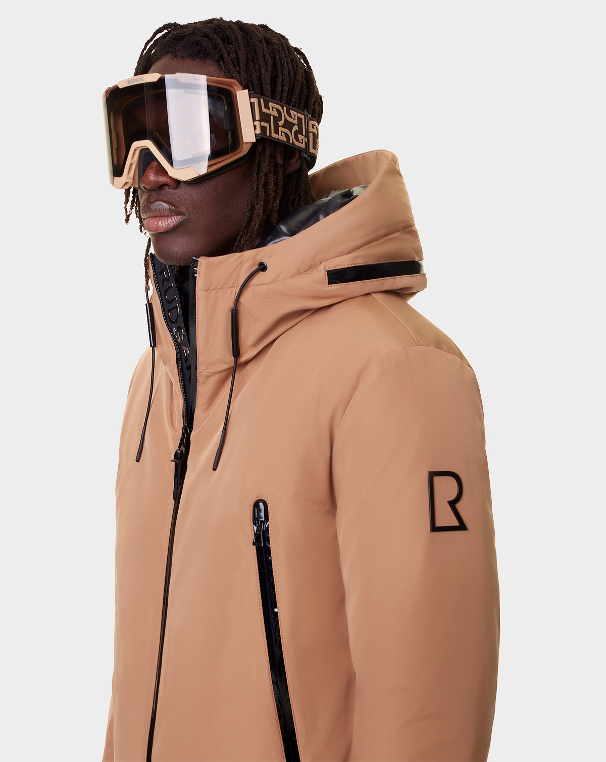 A person in a tan hooded winter jacket with black zipper accents and large Rudsak PEAK - 9421515 RUDSAK PATTERN ski goggles, featuring a bold geometric strap and interchangeable lenses, stands against a plain light background.