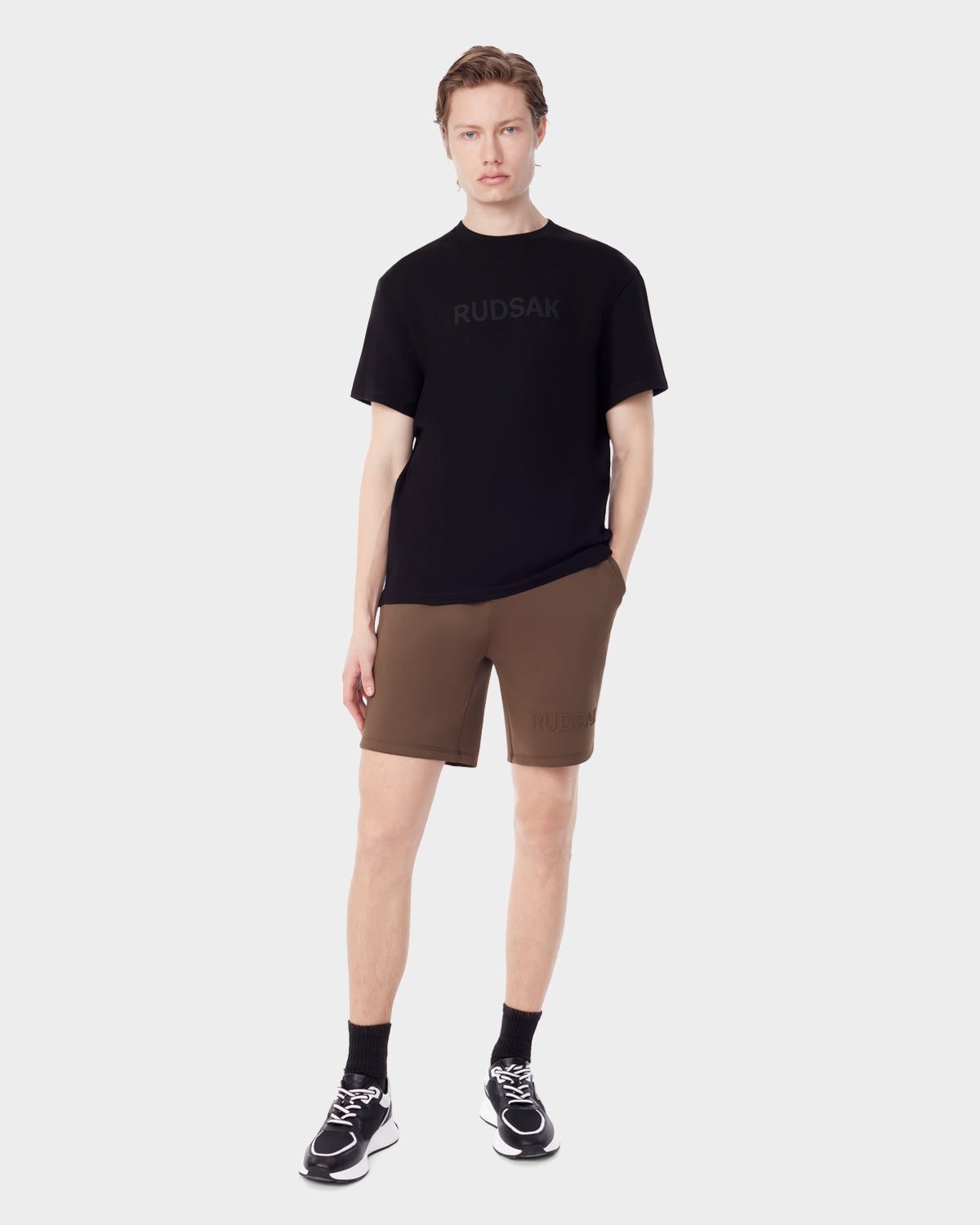 A person faces forward against a plain background wearing a black Rudsak t-shirt, ELLIS - 6523007 ARMY men's neoprene shorts by Rudsak, black socks, and black sneakers with white soles. One hand rests in their pocket.