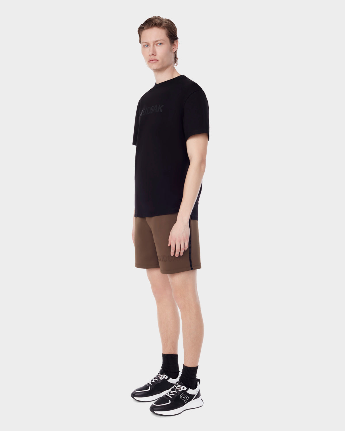 A young man stands against a white background wearing a black T-shirt, Rudsak ELLIS - 6523007 ARMY neoprene men's shorts, black socks, and black sneakers with white soles, facing slightly to the side with a neutral expression.