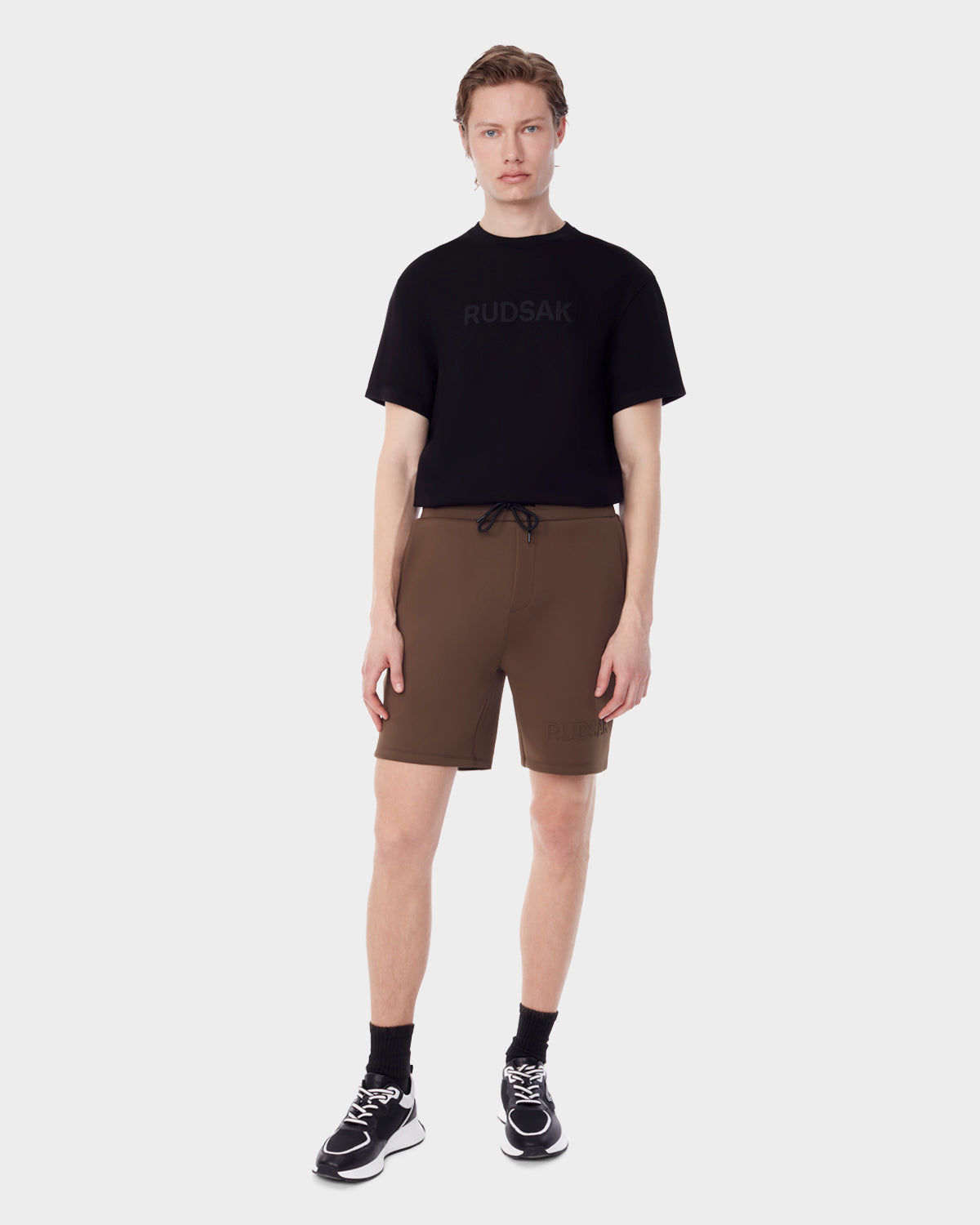 A person stands facing forward in a black Rudsak t-shirt, ELLIS - 6523007 ARMY neoprene men’s shorts in brown, black ankle socks, and black sneakers against a plain light background.