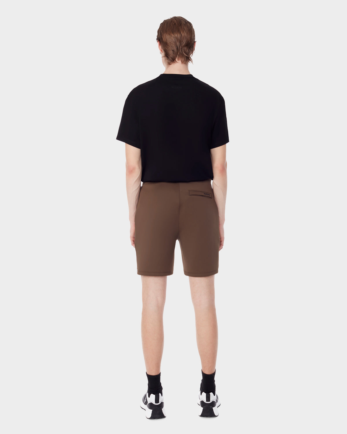 A person stands with their back to the camera, wearing a black t-shirt, Rudsak ELLIS - 6523007 ARMY men's neoprene shorts, black socks, and black-and-white sneakers against a plain light gray background.