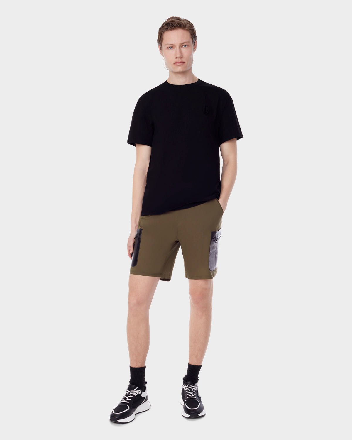 A person faces forward against a plain light background, wearing RUDSAK MICHAEL - 6523011 ARMY mens olive green stretch shorts with vegan leather cargo pockets, paired with a black t-shirt, black socks, and black-and-white sneakers. One hand is in their pocket.