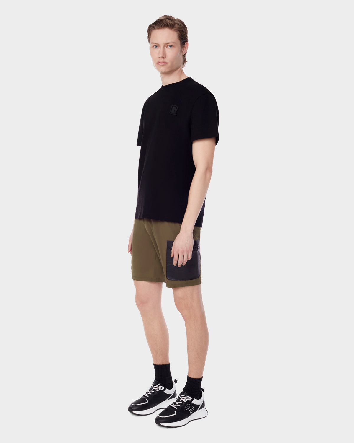 A man stands against a plain gray background, wearing RUDSAK’s MICHAEL - 6523011 ARMY mens shorts with black vegan leather cargo pockets, a black t-shirt, black socks, and black-and-white sneakers, looking forward with a neutral expression.