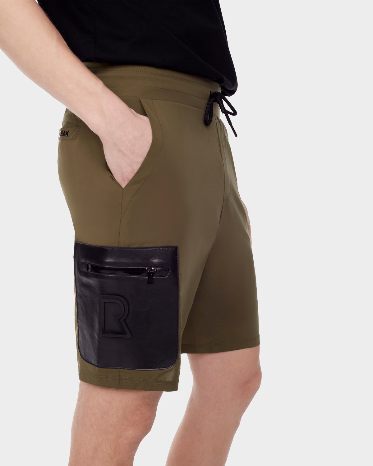 A person wears RUDSAKs MICHAEL - 6523011 ARMY mens olive green stretch-light shorts with a black vegan leather zip pocket and embossed R detail, paired with a black T-shirt, against a plain light background.