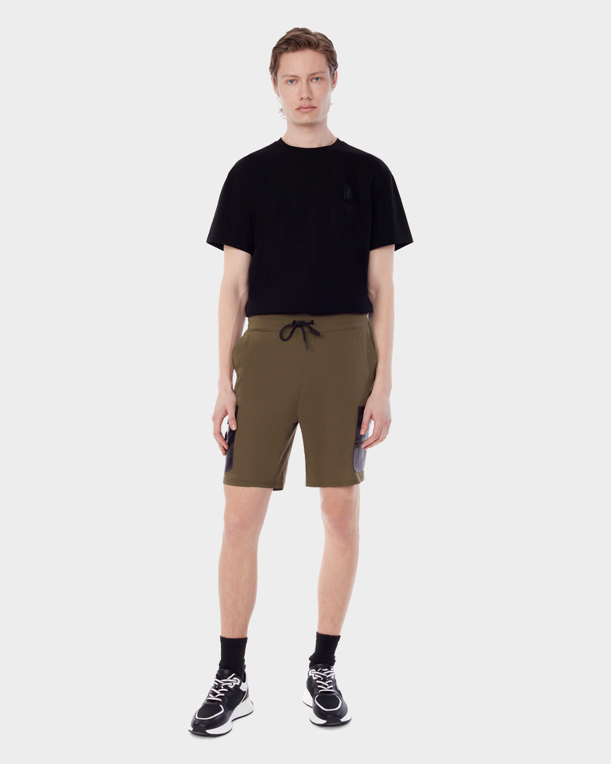 A young man stands facing forward, wearing RUDSAK MICHAEL - 6523011 ARMY mens shorts with black side panels, a black t-shirt, black socks, and black sneakers with white soles against a plain light background.