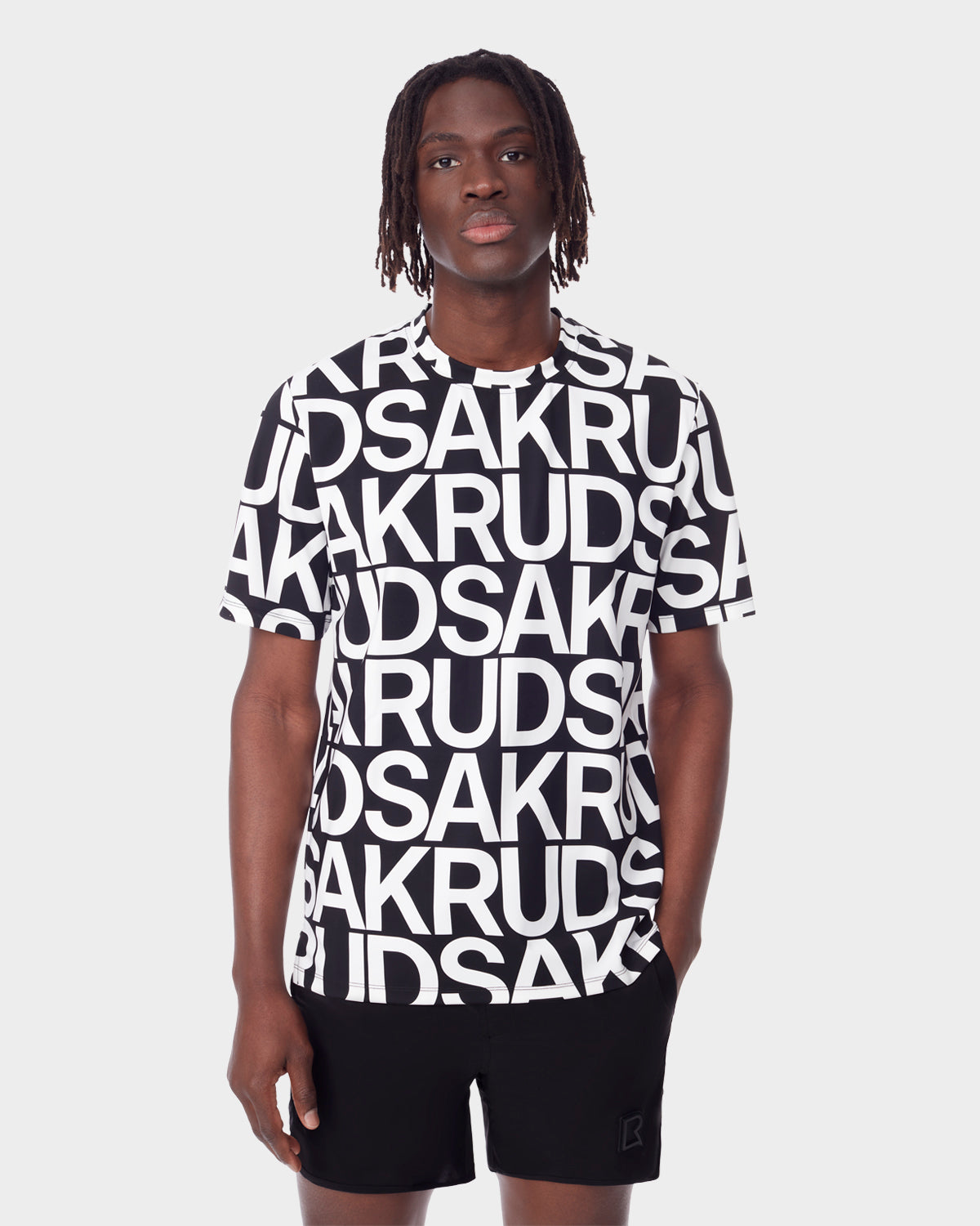 A man with medium-length dreadlocks wears the RAYA - 9523004 M BLACK ALPHABET unisex T-shirt by Rudsak, featuring bold black and white letters, paired with black shorts, standing against a plain white background.