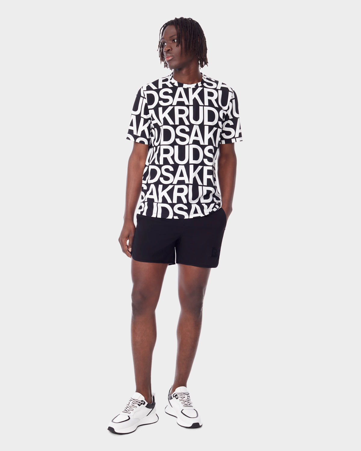 A man models the Rudsak RAYA - 9523004 M BLACK ALPHABET unisex T-shirt with bold black-and-white graphic letters, paired with black shorts and white sneakers. He stands against a plain background, looking slightly left with relaxed hands at his sides.