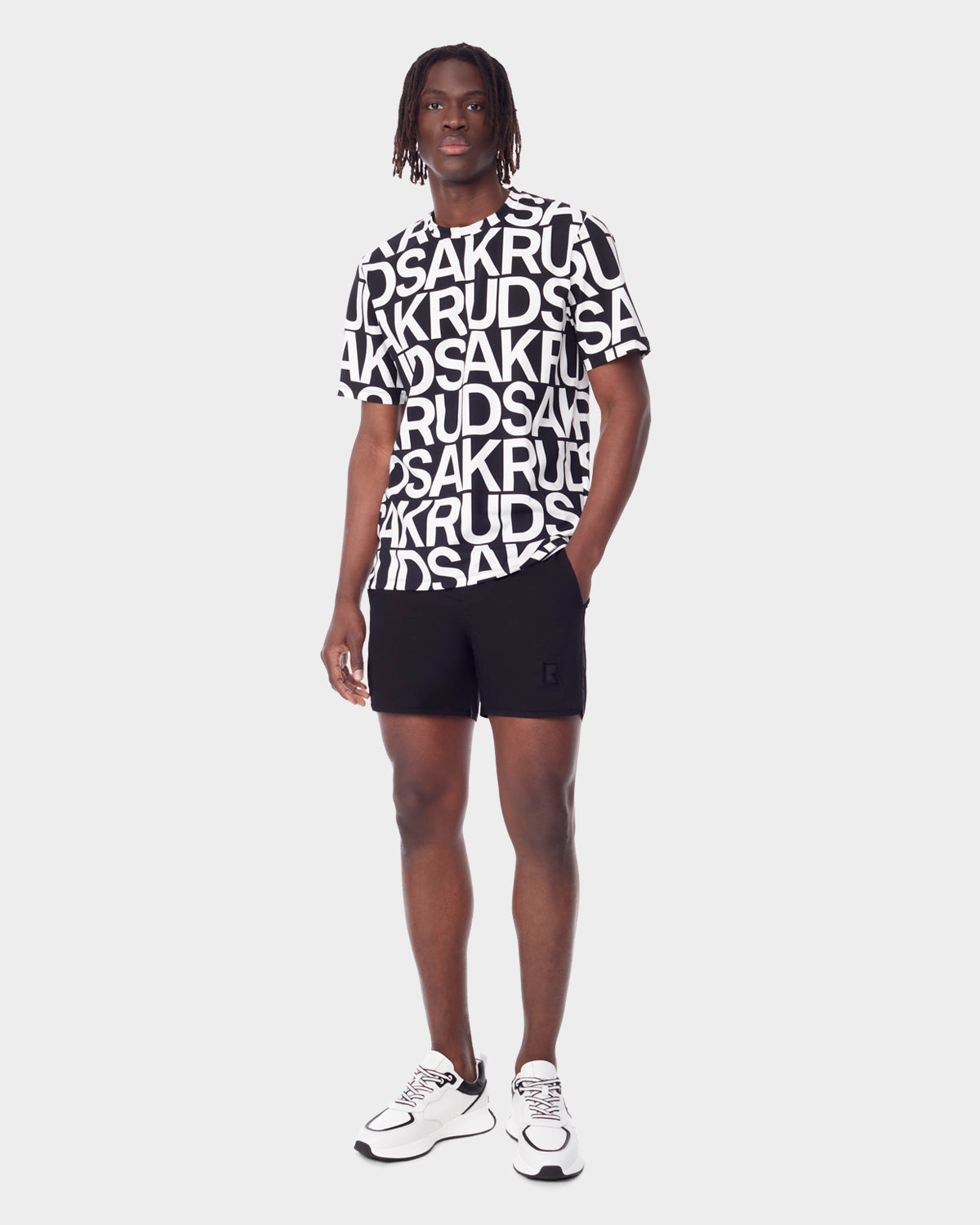 A man with short, twisted hair and a neutral expression wears the Rudsak RAYA - 9523004 M BLACK ALPHABET unisex T-shirt, featuring bold overlapping letters, paired with black shorts and white sneakers against a plain background.