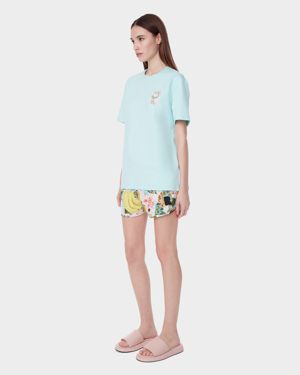 A woman with straight brown hair and a neutral expression stands against a white background, wearing the Rudsak DIAR - 9523003 W ICE BLUE t-shirt with a small chest graphic and unisex fit, paired with colorful shorts and pale pink sandals.