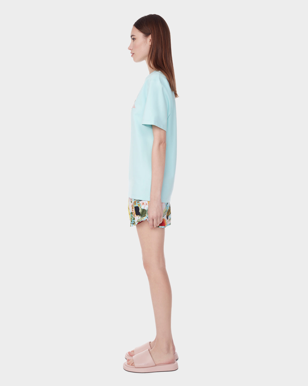 A woman stands in profile against a white background, wearing the Rudsak DIAR - 9523003 W ICE BLUE unisex-fit light blue t-shirt, floral shorts, and light pink slide sandals.