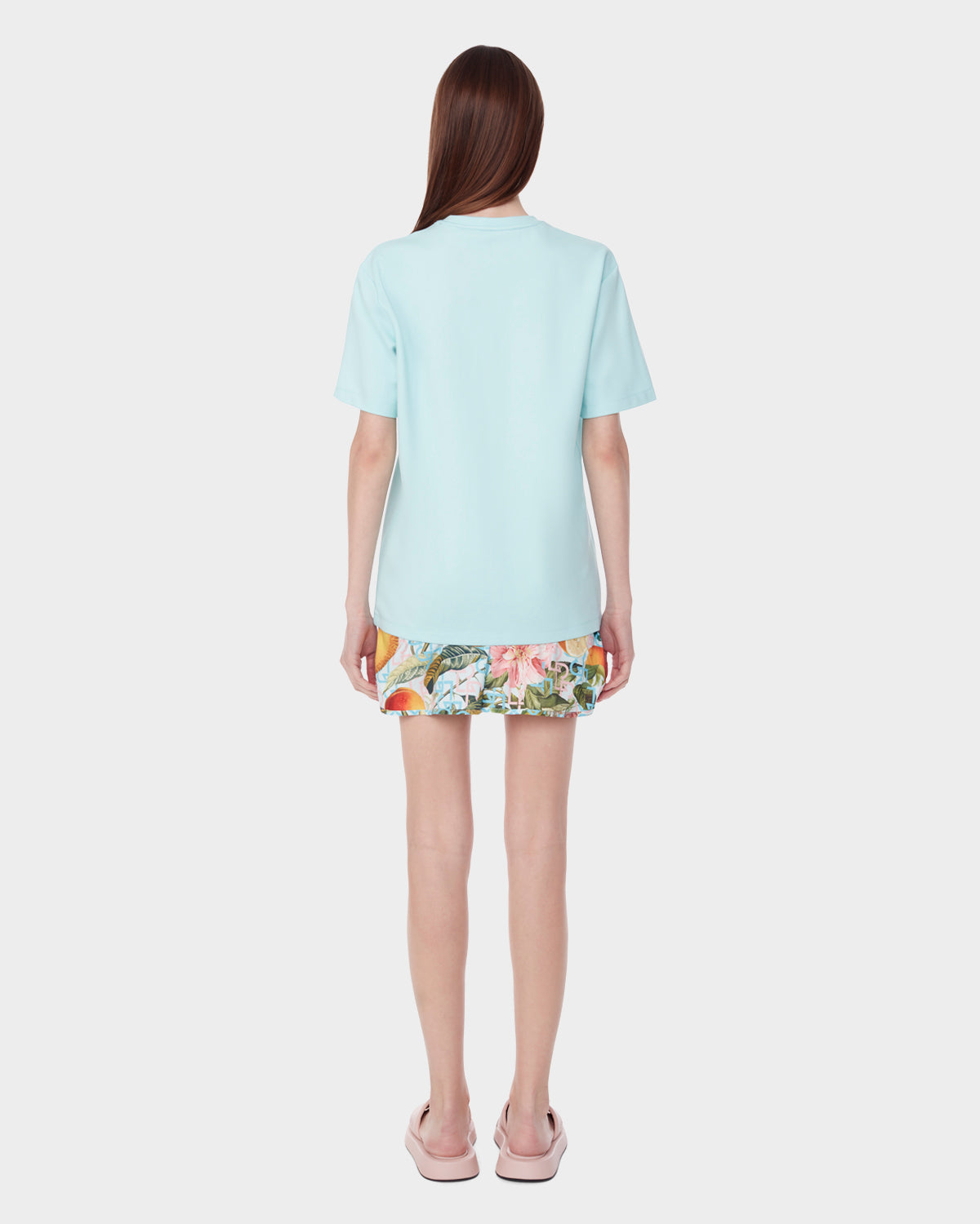 A woman faces away, wearing the Rudsak DIAR - 9523003 W ICE BLUE short-sleeve shirt, a colorful floral skirt, and light pink slide sandals against a plain white background.