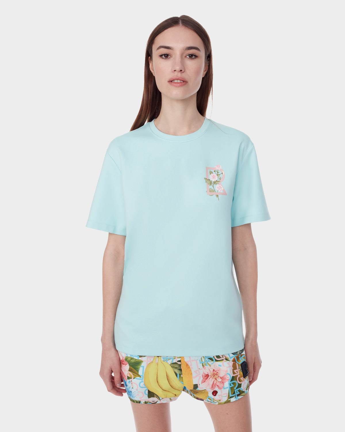 A woman with straight brown hair models the Rudsak DIAR - 9523003 W ICE BLUE T-shirt against a white background, paired with vibrant fruit-patterned shorts.