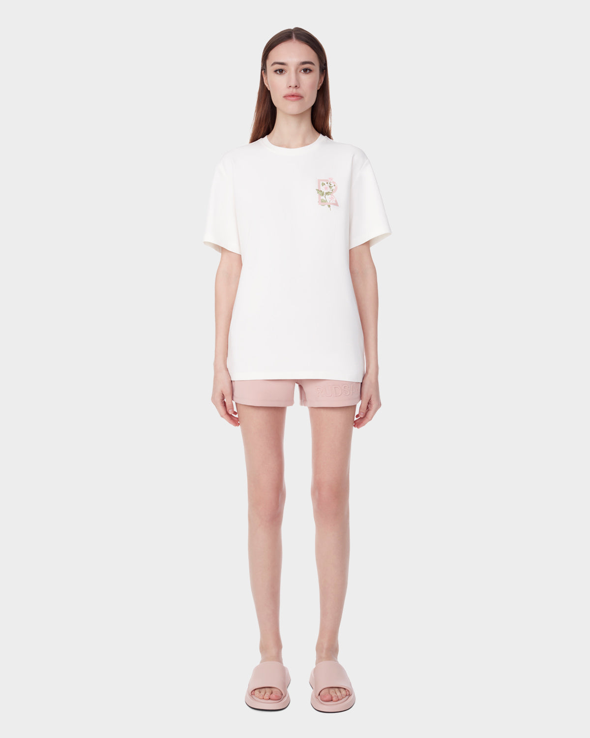 A woman stands facing forward against a plain light background, wearing the Rudsak DIAR - 9523003 W OPTIC WHITE T-shirt featuring a small floral chest design, pale pink shorts, and matching pink slide sandals.