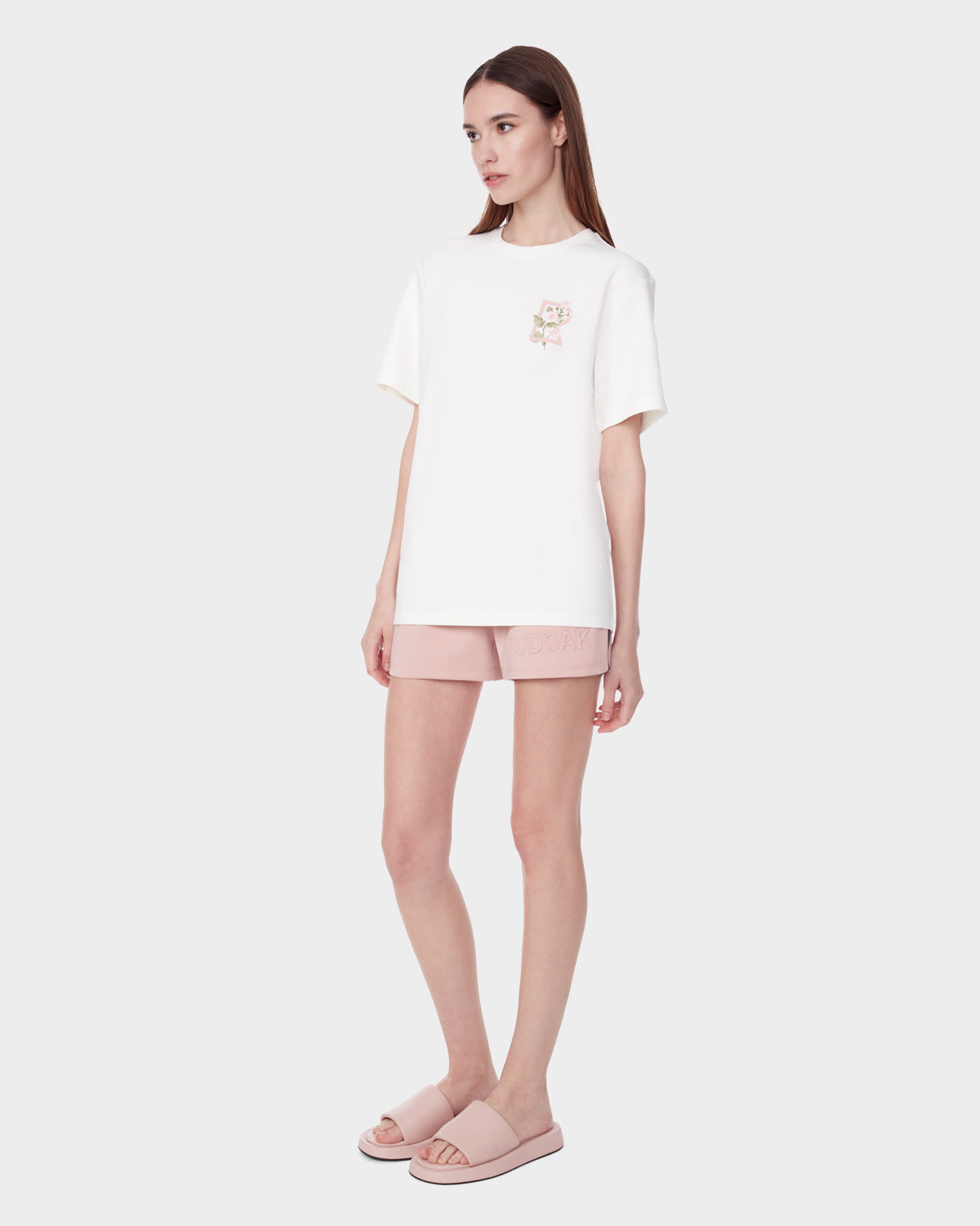A woman with straight brown hair and a neutral expression models the Rudsak DIAR - 9523003 W OPTIC WHITE T-shirt, featuring a small floral chest graphic, paired with light pink shorts and matching slide sandals against a plain background.