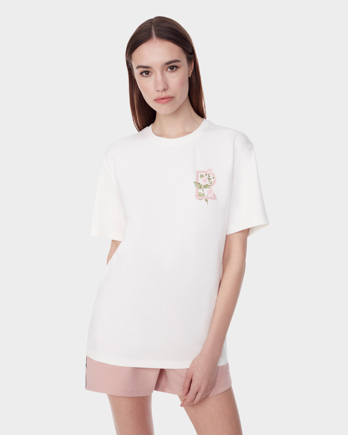 A woman with straight brown hair wears the Rudsak DIAR - 9523003 W OPTIC WHITE short-sleeve T-shirt, featuring a small floral chest overlay, paired with pale pink shorts against a plain light background.