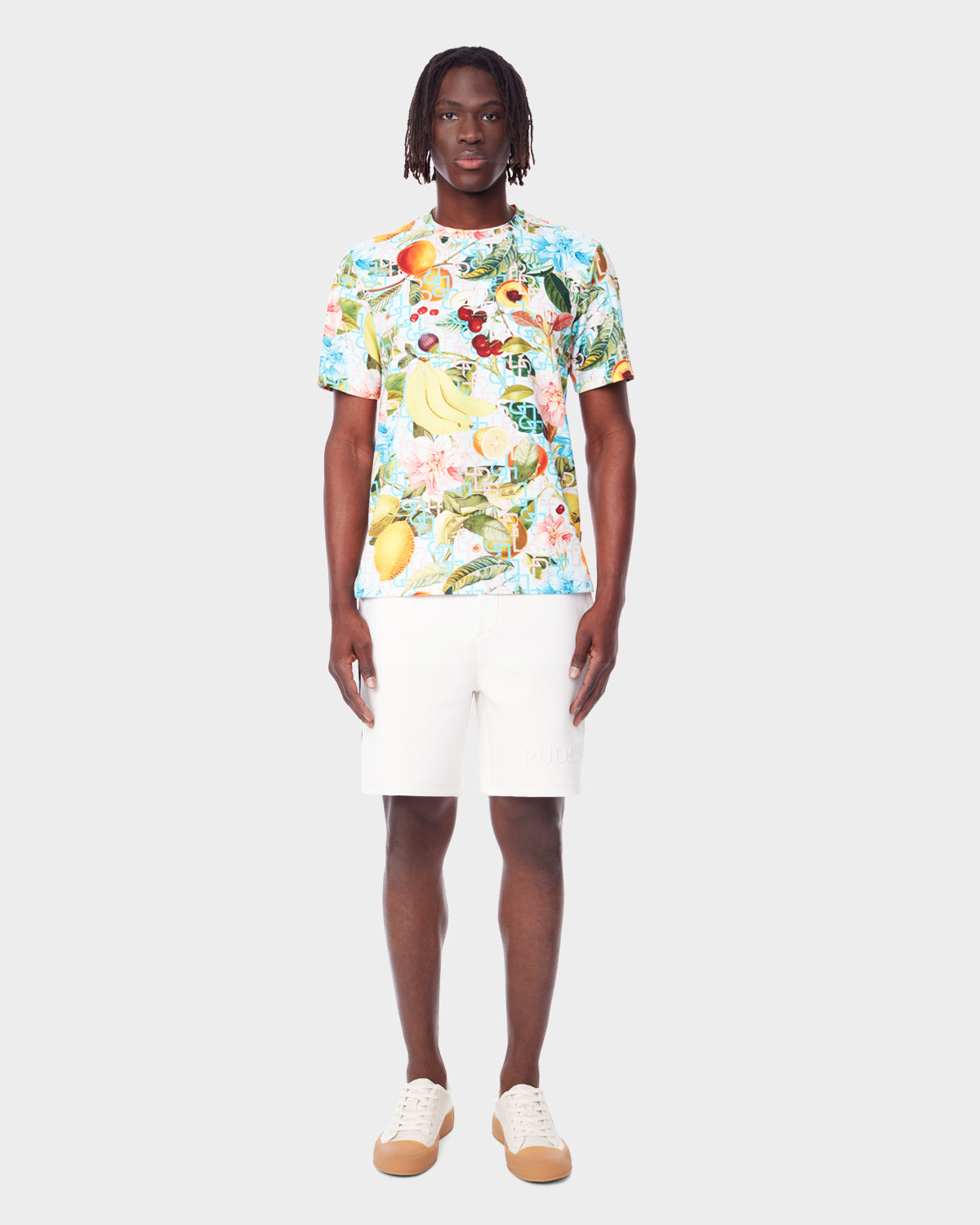A man stands facing forward, wearing the RAYA - 9523004 COCKTAIL RUDSAK M unisex T-shirt by Rudsak, paired with white shorts and white sneakers with tan soles, against a plain light gray background.