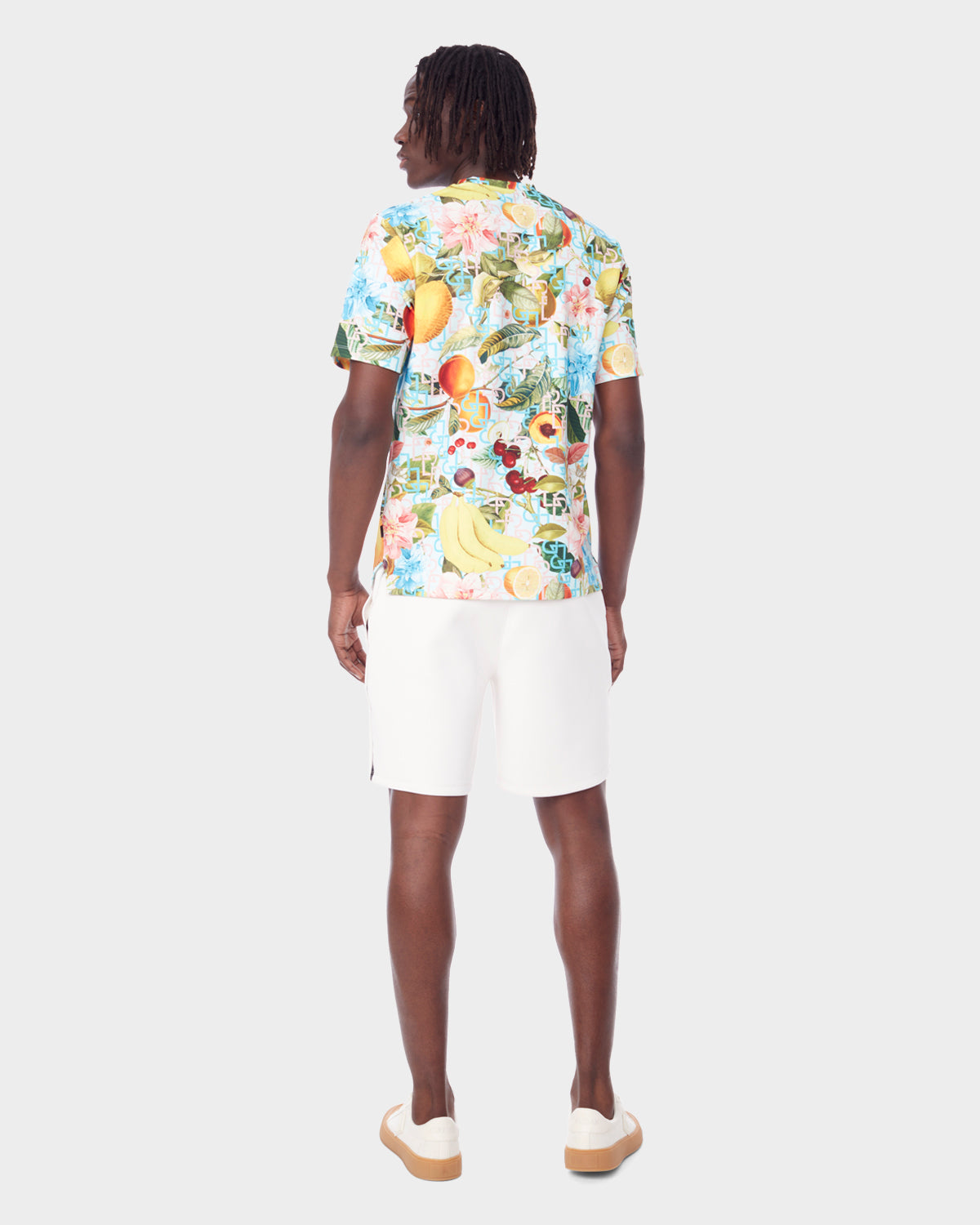 A man faces away wearing the Rudsak RAYA - 9523004 COCKTAIL RUDSAK M unisex fruit cocktail print T-shirt, paired with white shorts and sneakers, set against a plain light gray background.