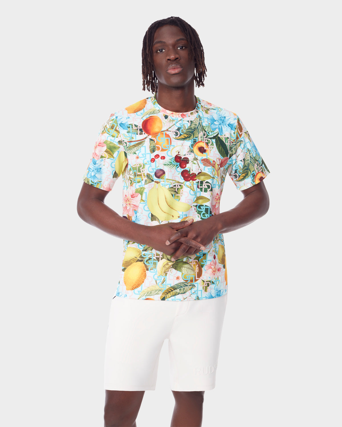 A man faces forward, hands loosely clasped, in a Rudsak RAYA - 9523004 COCKTAIL RUDSAK M unisex T-shirt with a vibrant fruit cocktail print and plain white shorts, set against a simple background.