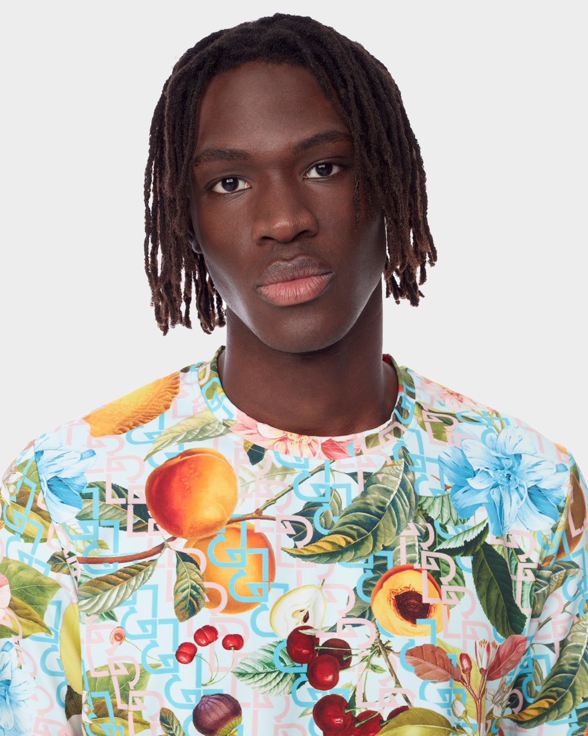 A young man with medium-length dreadlocks models the Rudsak RAYA - 9523004 COCKTAIL RUDSAK M unisex T-shirt, featuring a vibrant fruit cocktail and floral print, standing against a plain white background with a neutral expression.