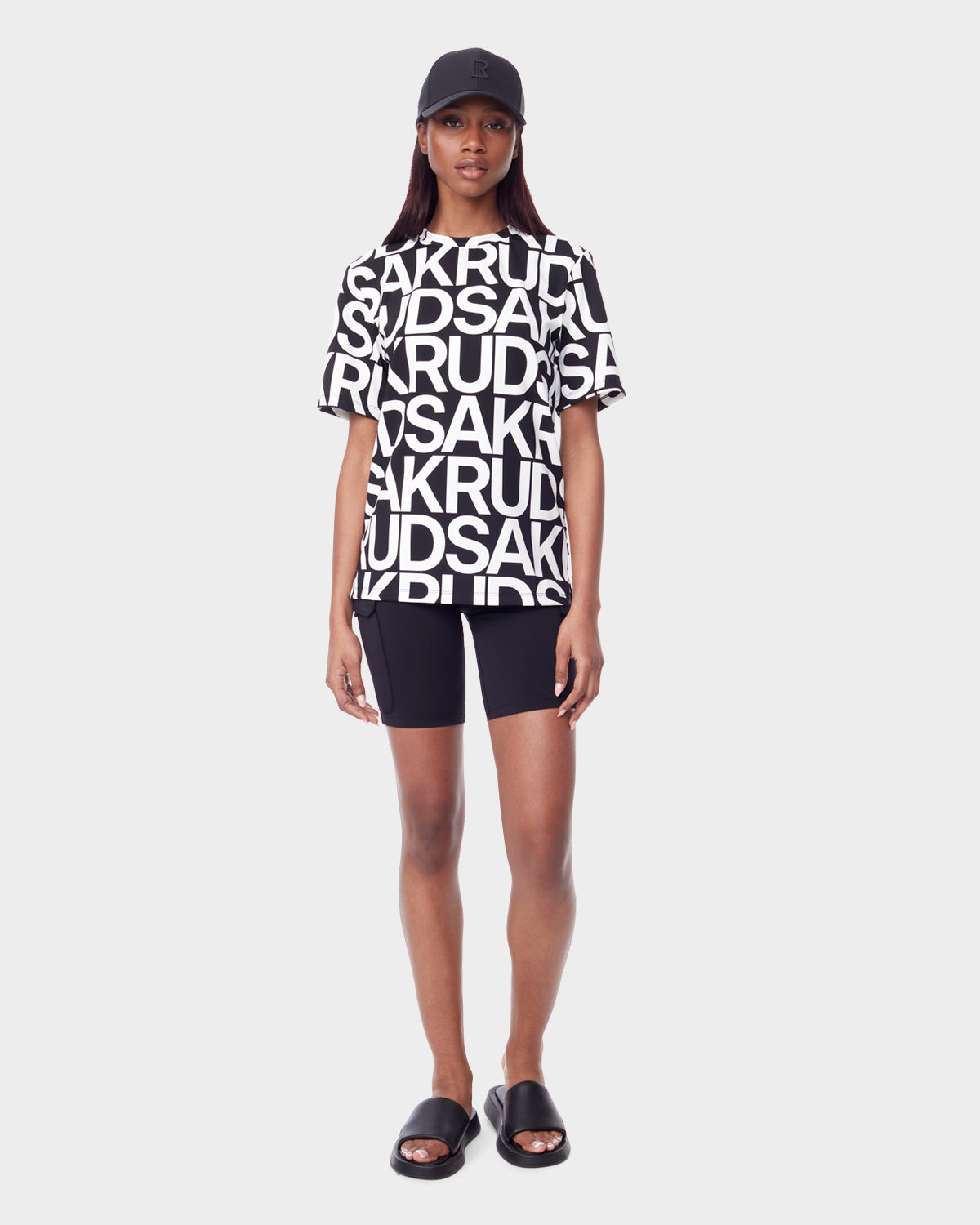 A woman stands against a plain background wearing the Rudsak RAYA - 9523004 W BLACK ALPHABET color-blocked unisex T-shirt with bold letters, paired with a black cap, black biker shorts, and slide sandals.