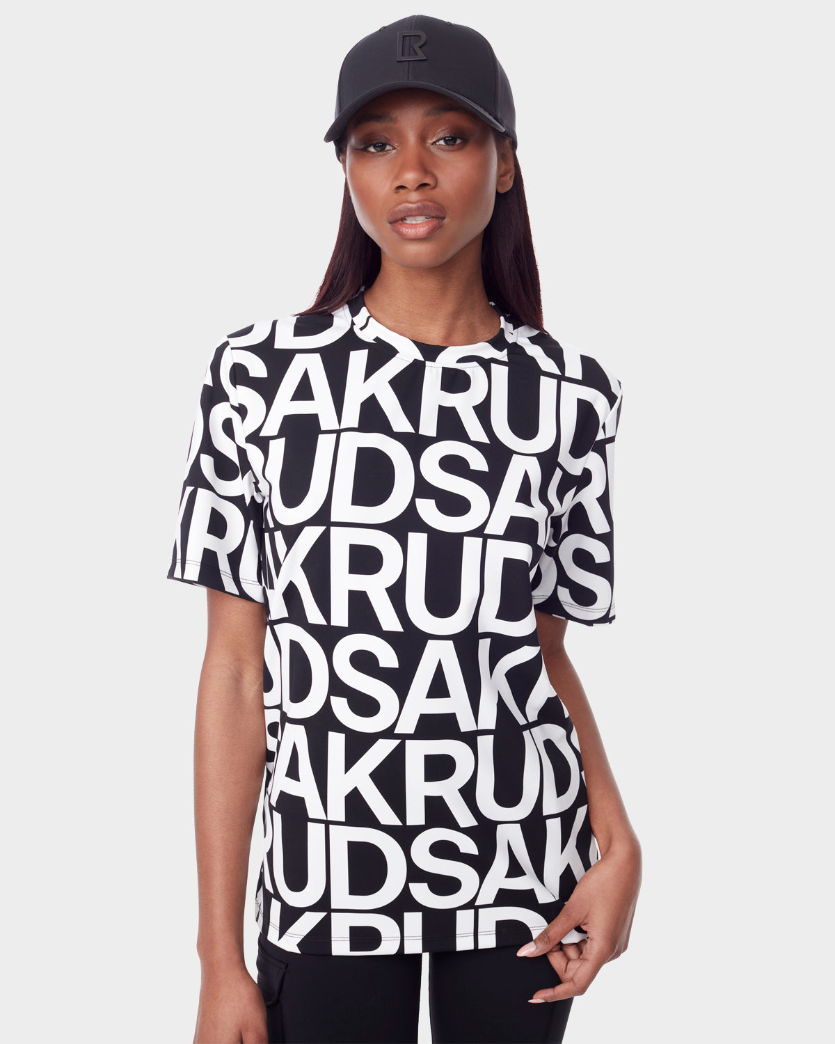 A woman in a black cap and the Rudsak RAYA - 9523004 W BLACK ALPHABET unisex T-shirt, featuring bold overlapping "RUDSAK" letters in black and white, stands against a plain light background.