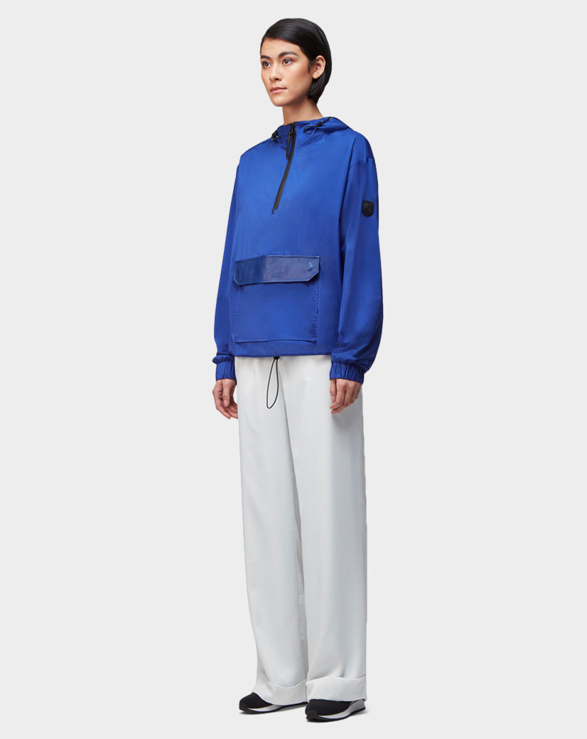 A person stands facing forward wearing the Rudsak STALENS - CA EN 9520000 ROYAL BLUE unisex pullover hoodie windbreaker with a front pocket, paired with loose white pants and black shoes against a plain light background.