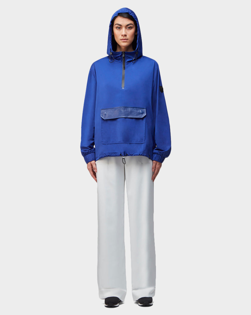 A person stands facing forward, wearing the Rudsak STALENS - CA EN 9520000 ROYAL BLUE hoodie windbreaker pullover with a front zip and large pocket, paired with loose white pants and black shoes, against a plain light gray background.