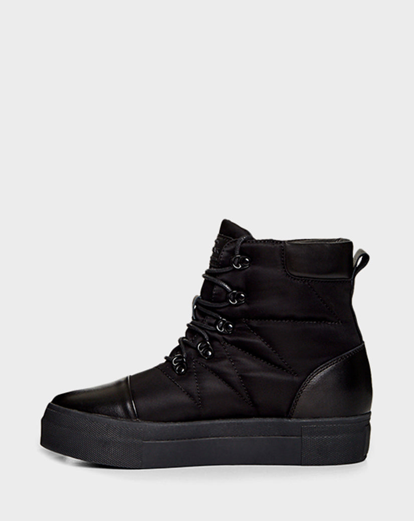 The Rudsak TASSEL - CA EN 8219514 BLACK is a black high-top sneaker boot with a thick platform sole, metal eyelets, lace-up front, quilted side stitching, and smooth toe cap—ideal for winter style. Shown on a light gray background.